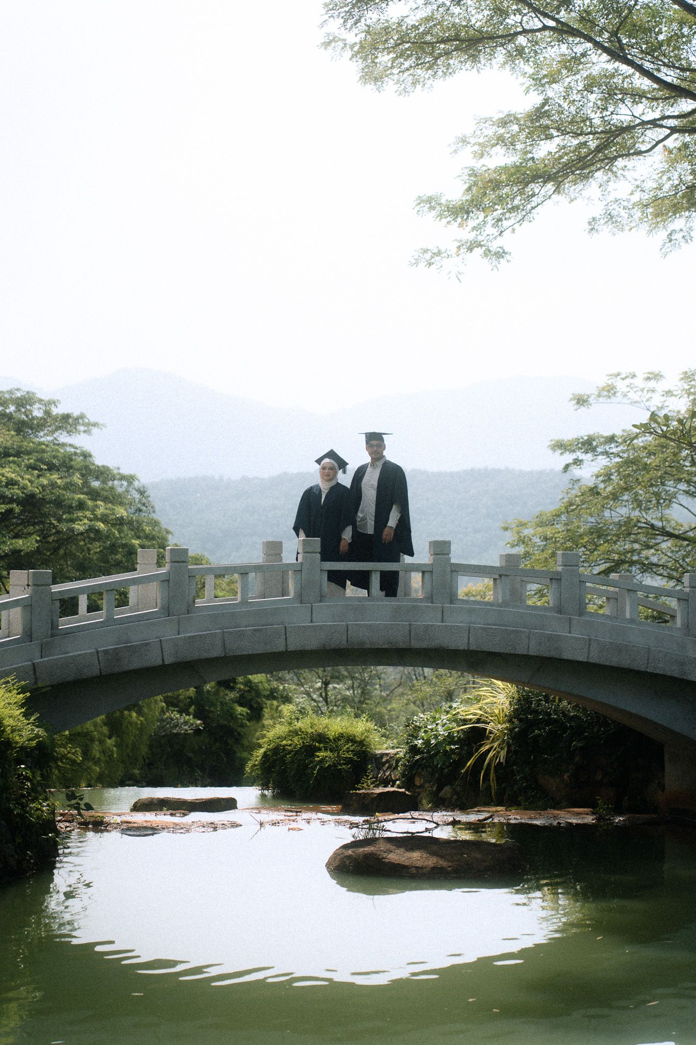 Two graduates in caps and gowns pose on a stone arch bridge over a serene pond surrounded by lush greenery.