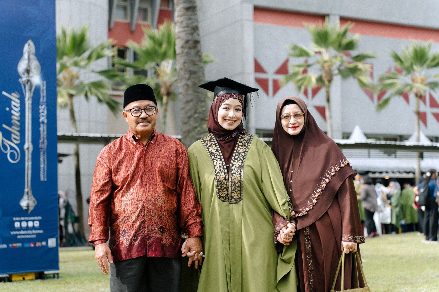 A graduate in cap and green gown poses with two family members at an outdoor university graduation ceremony.