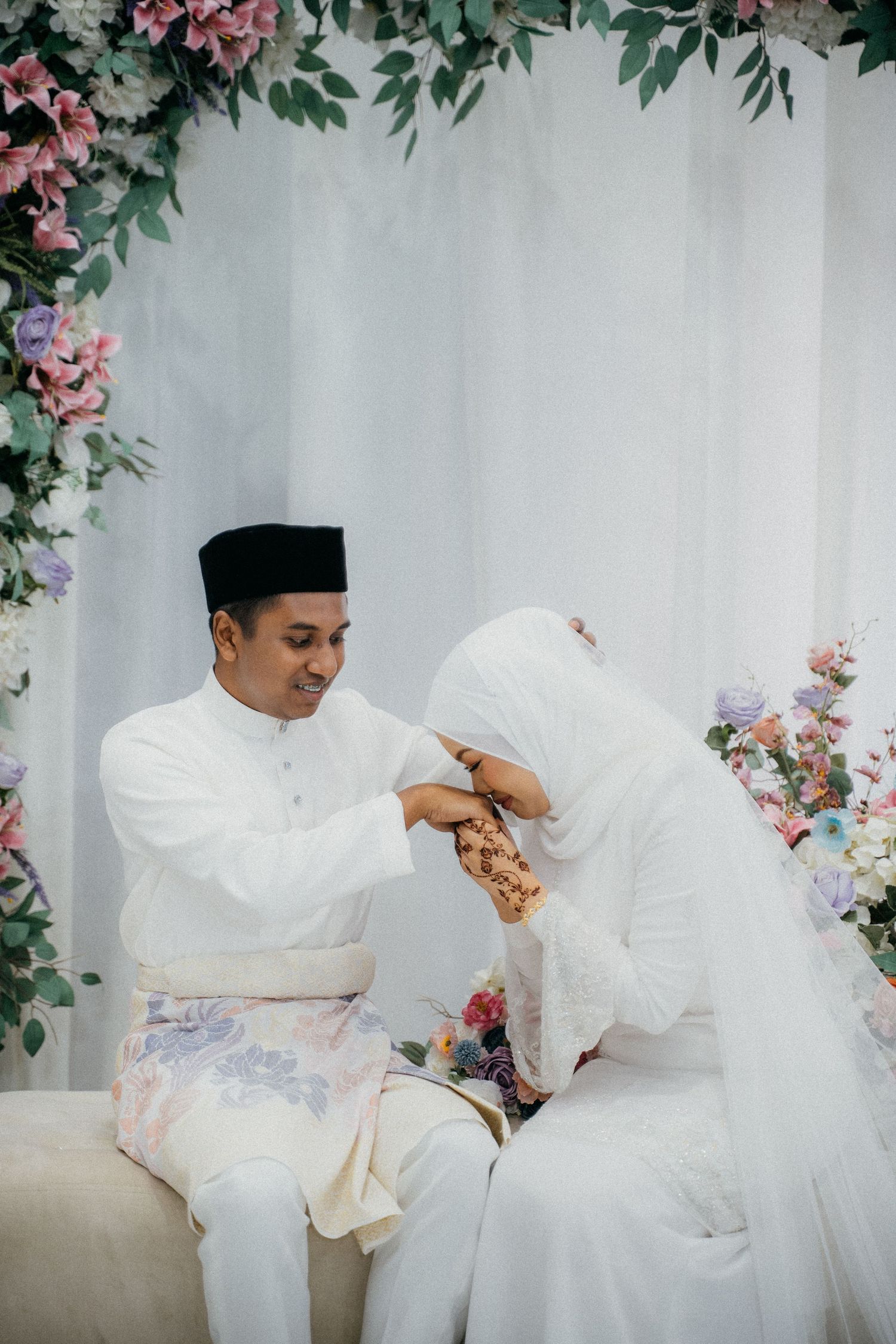 Traditional Malay wedding scene with flowing white outfits amid pink floral decorations against a light backdrop.