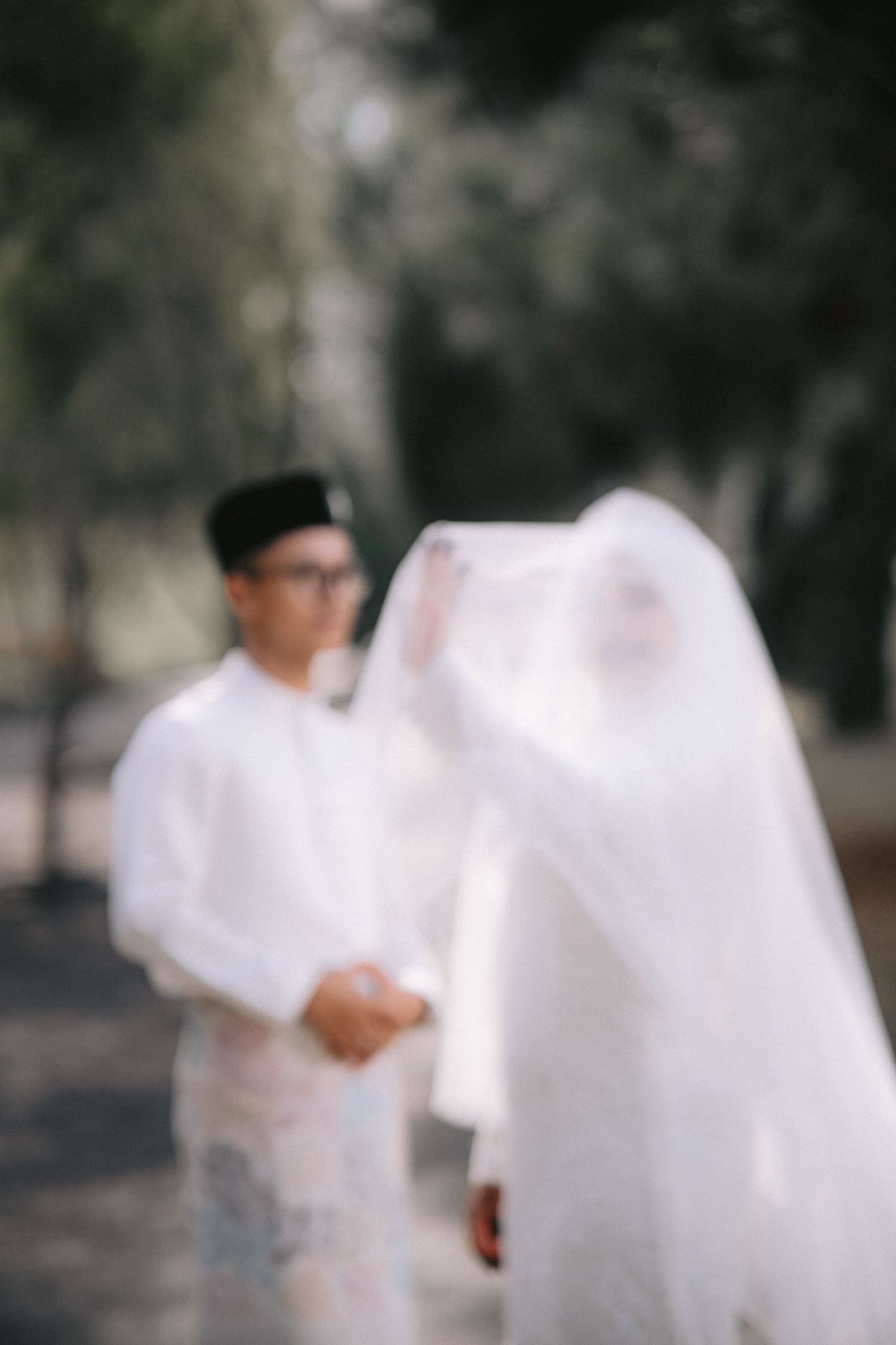 A blurred artistic wedding photoshoot shows soft silhouettes of a couple in white against a dark outdoor background.