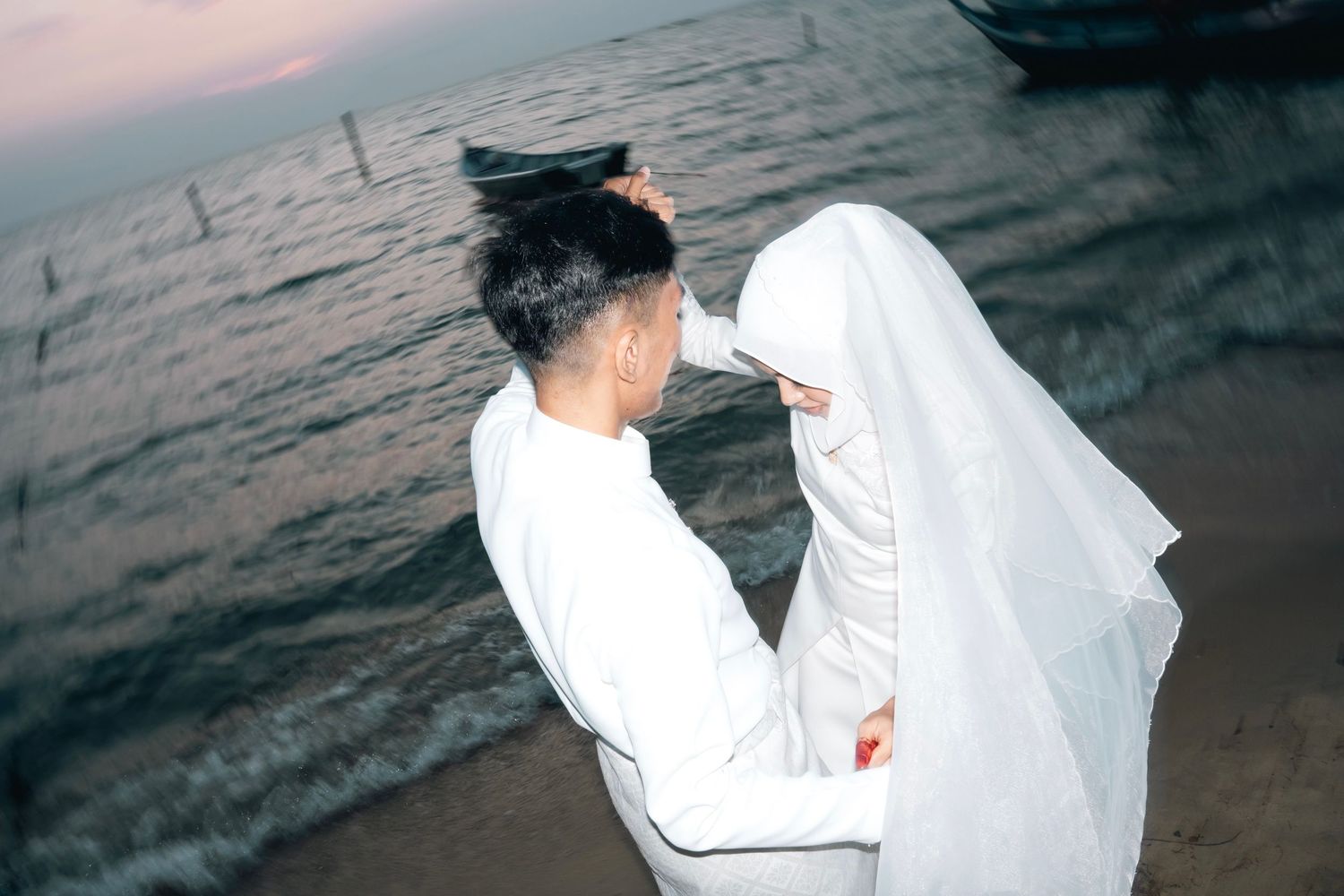 A romantic couple in white attire embraces on a beach at sunset with flowing veil in the ocean breeze.