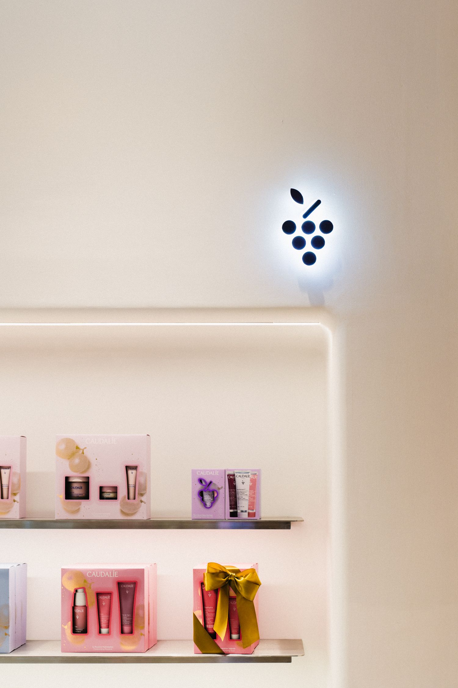 Minimalist wall display with glowing grape logo and pink beauty products on floating shelves.