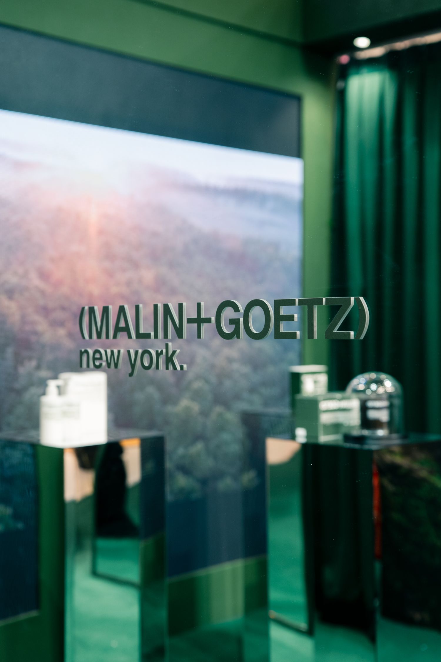 Blurred storefront sign showing Malin+Goetz New York branding in green tones.