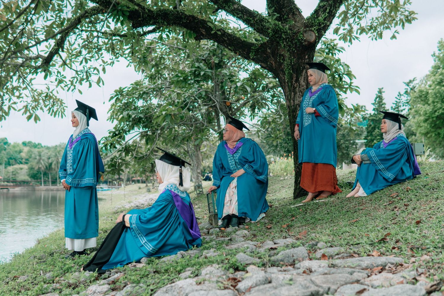 Graduates in blue gowns relaxing under a tree by a scenic lake.