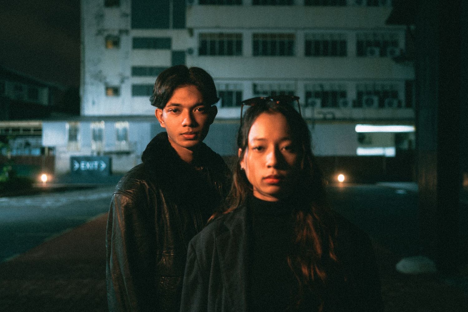 A moody nighttime urban portrait showing two figures in dark clothing standing close together.