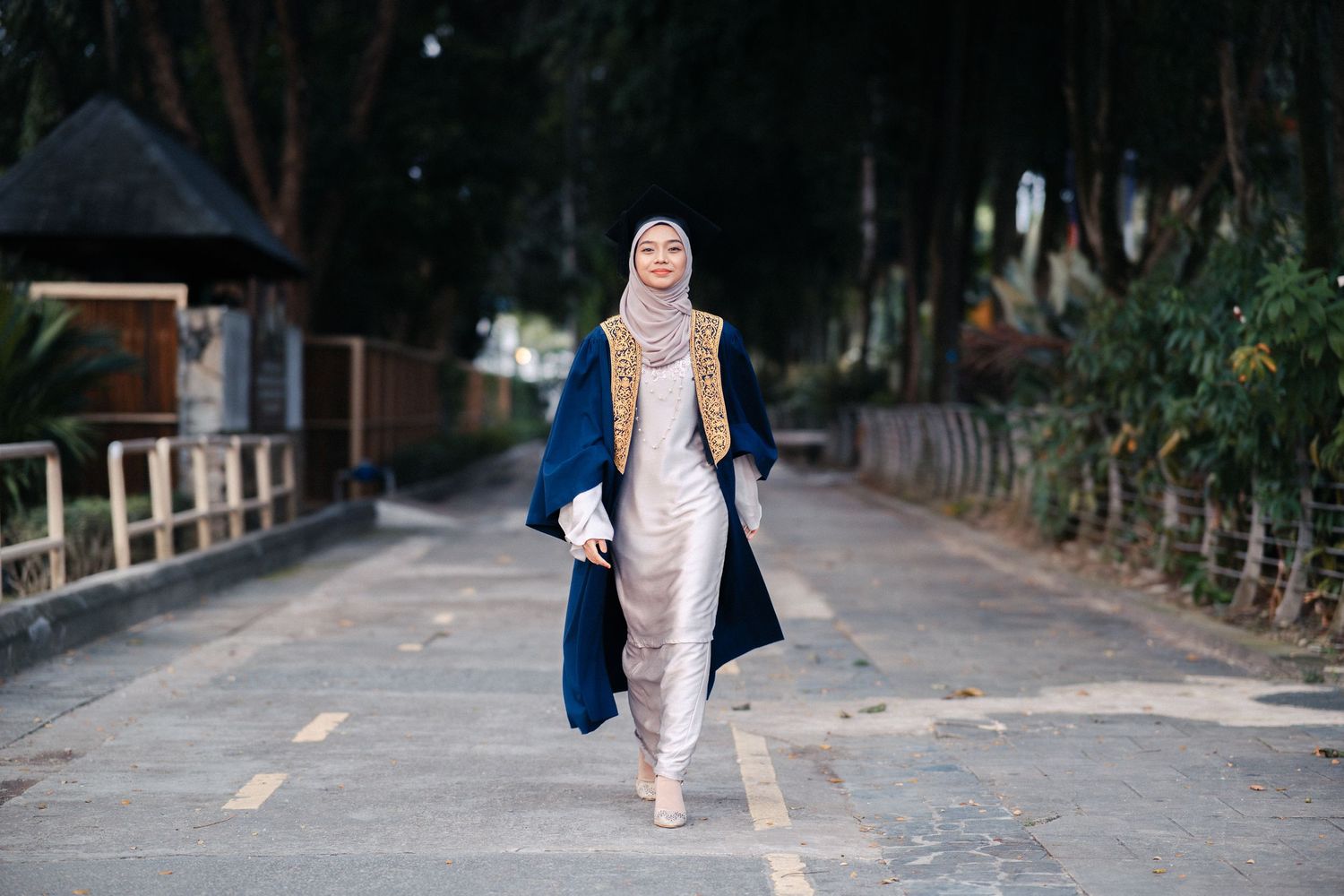 Graduate in blue gown walking along tree-lined path.