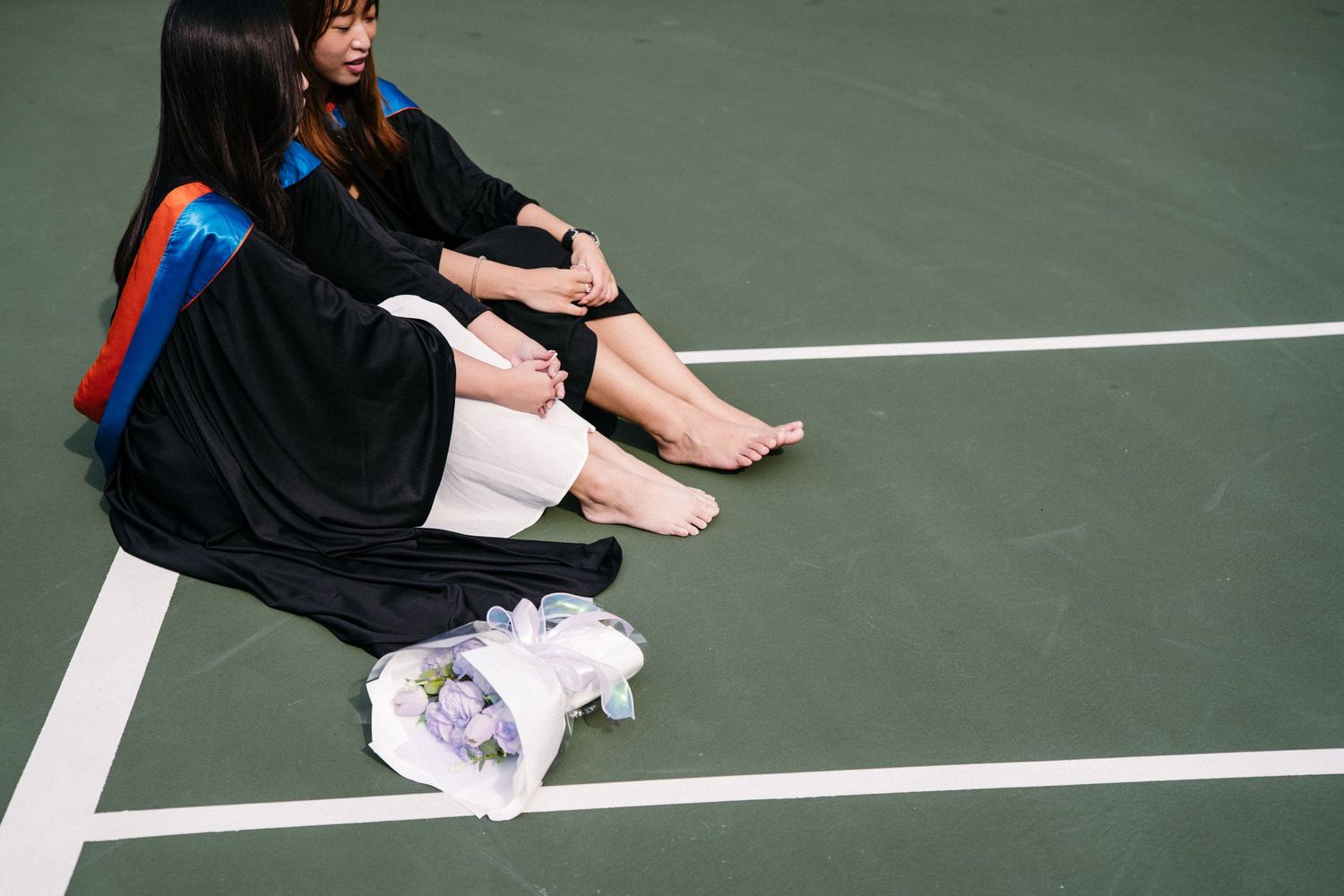 Students in graduation gowns resting on a tennis court with backpacks and belongings.