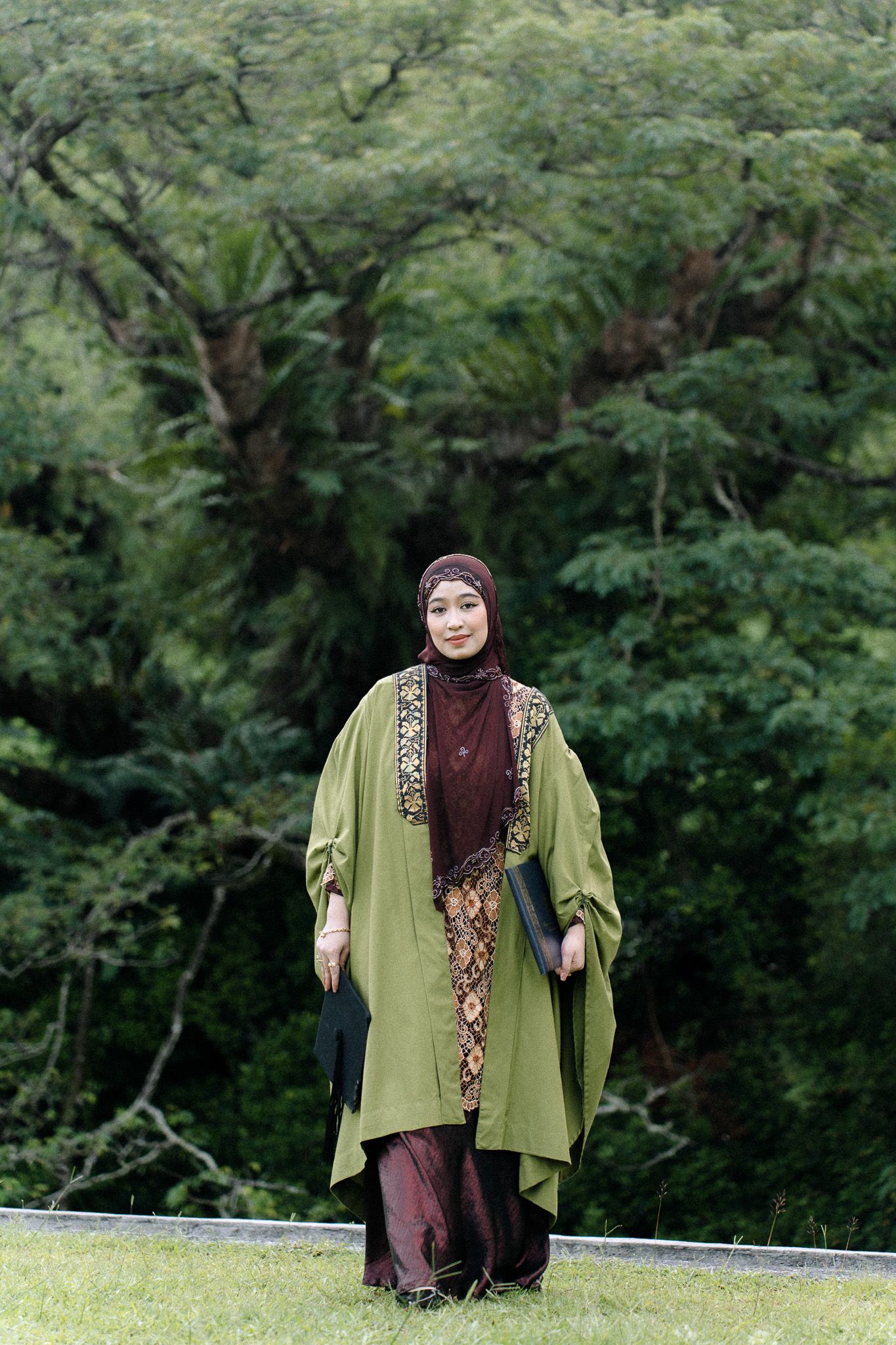 Muslim woman in green abaya and brown hijab stands on grass with lush green trees in background.