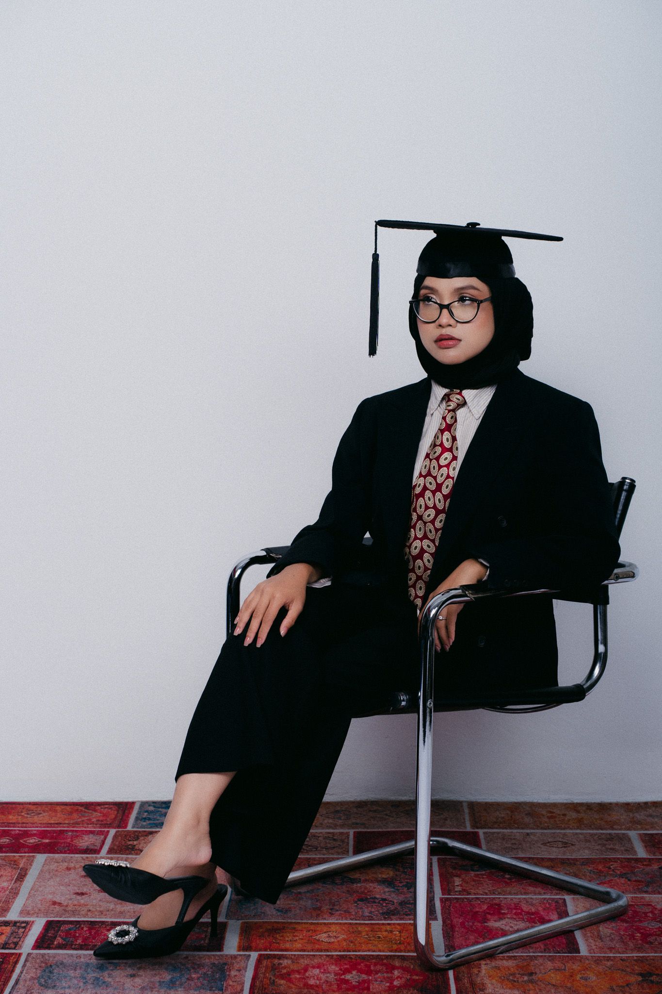 Young woman in black suit, graduation cap, hijab, glasses, and heels sits confidently on a chair against a white wall.