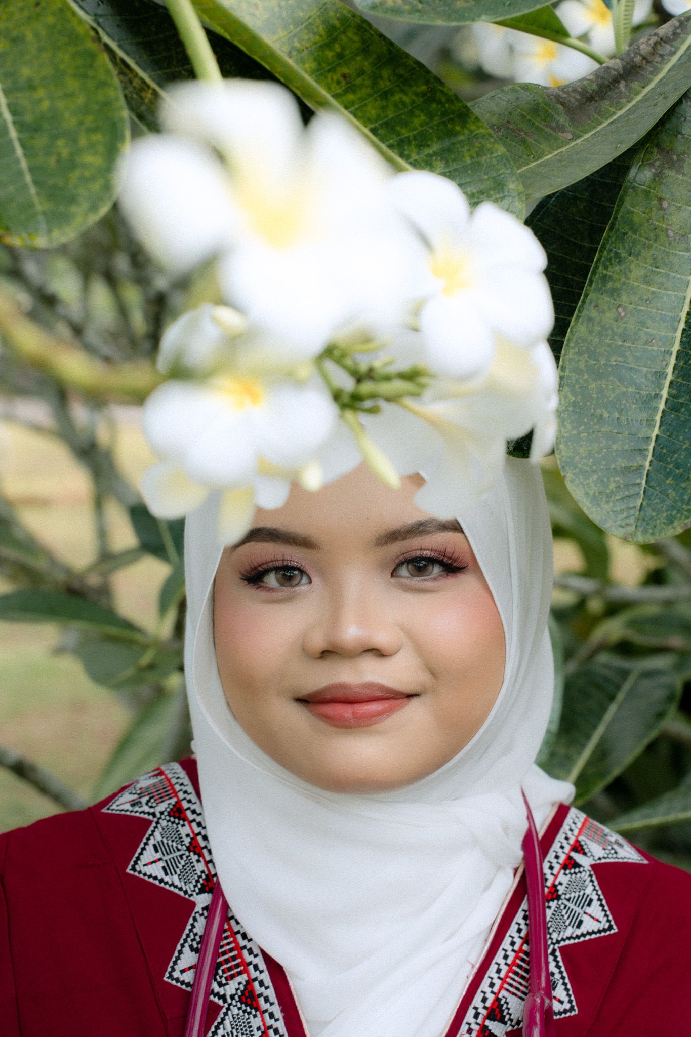 Young woman in white hijab and red embroidered top smiles beneath white plumeria flowers outdoors.