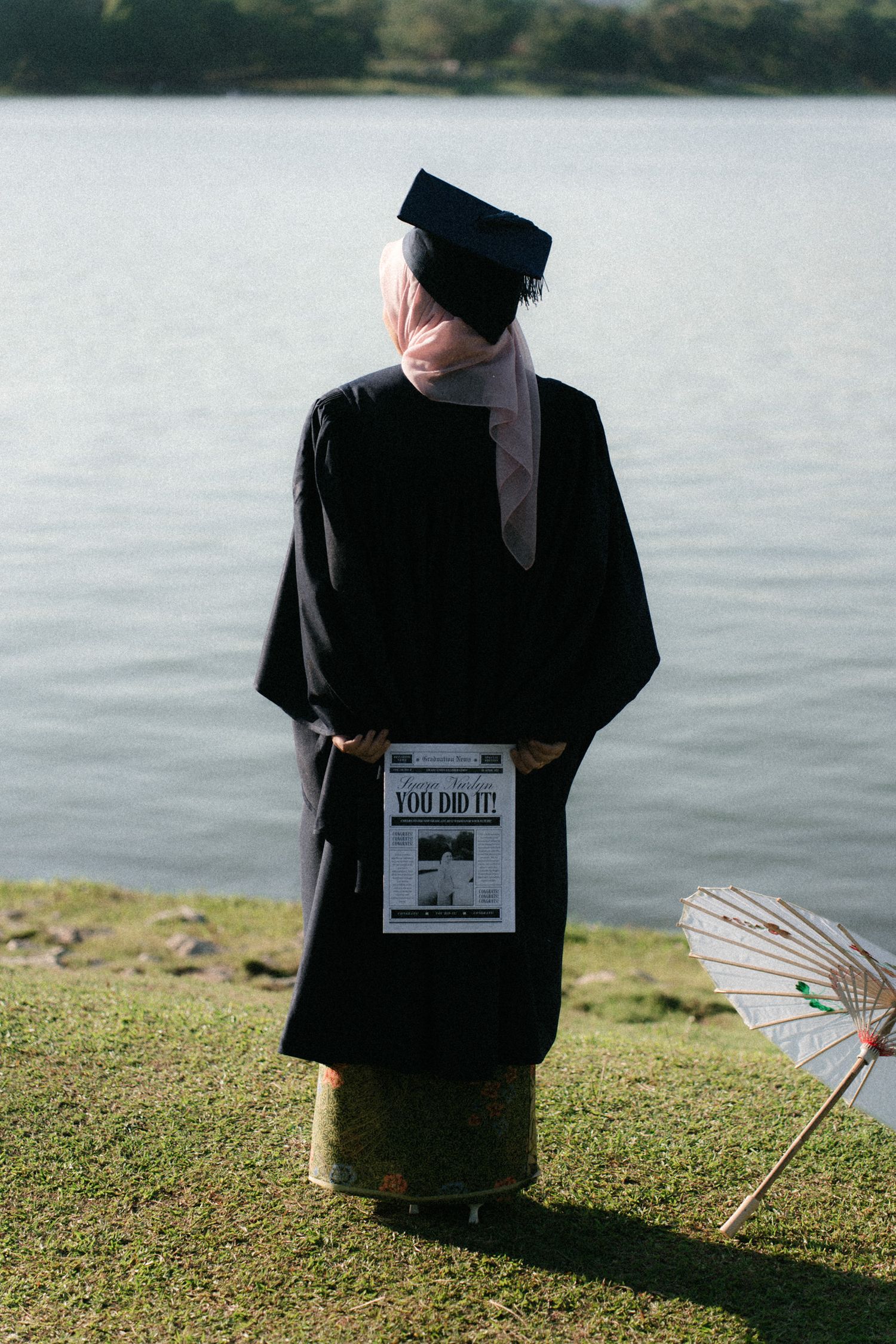 Graduate in black academic regalia sits by a lake with a decorative fan during a graduation photoshoot.