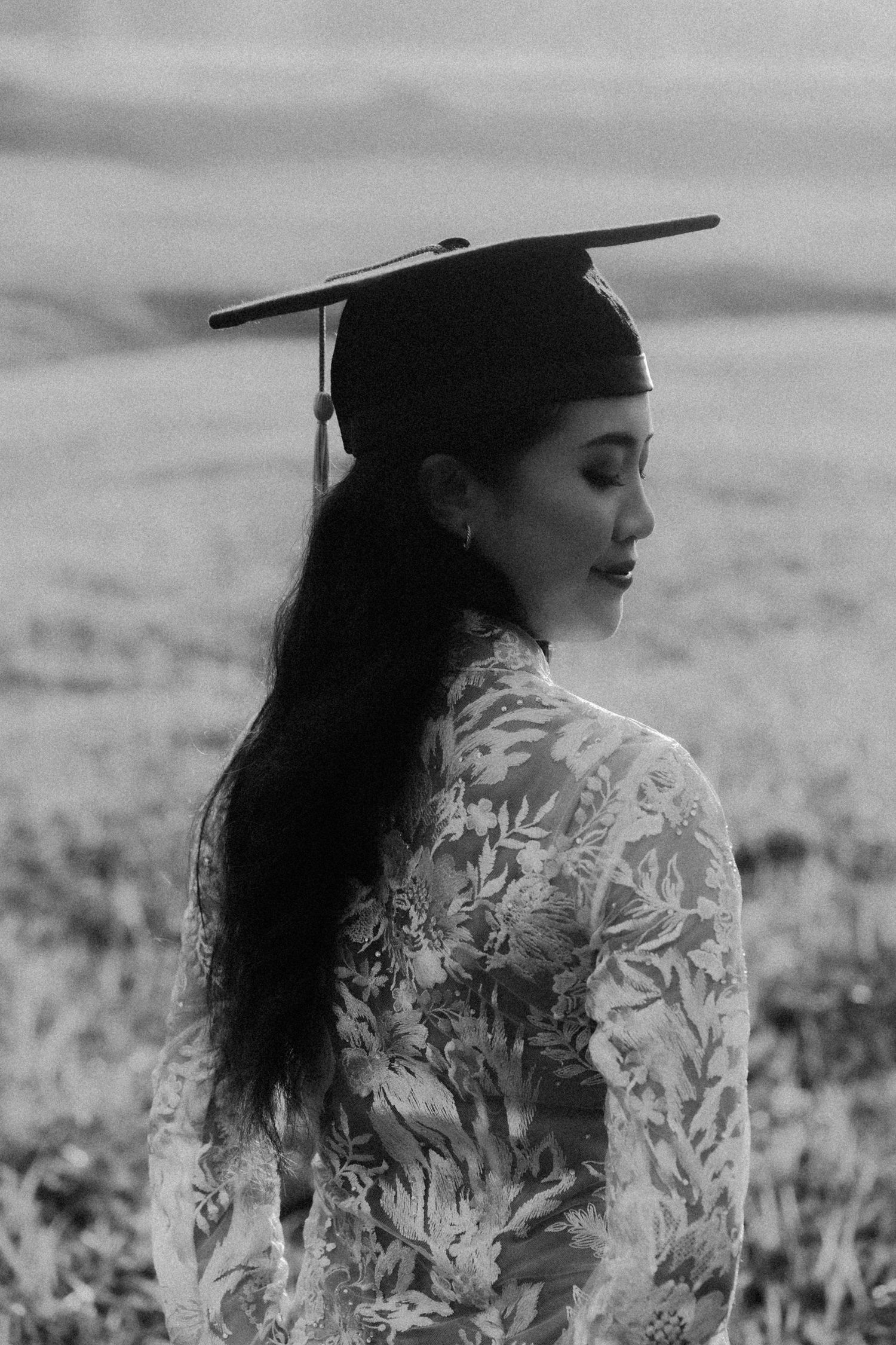 Black and white portrait of a graduate in lace dress and cap against a dreamy outdoor backdrop.
