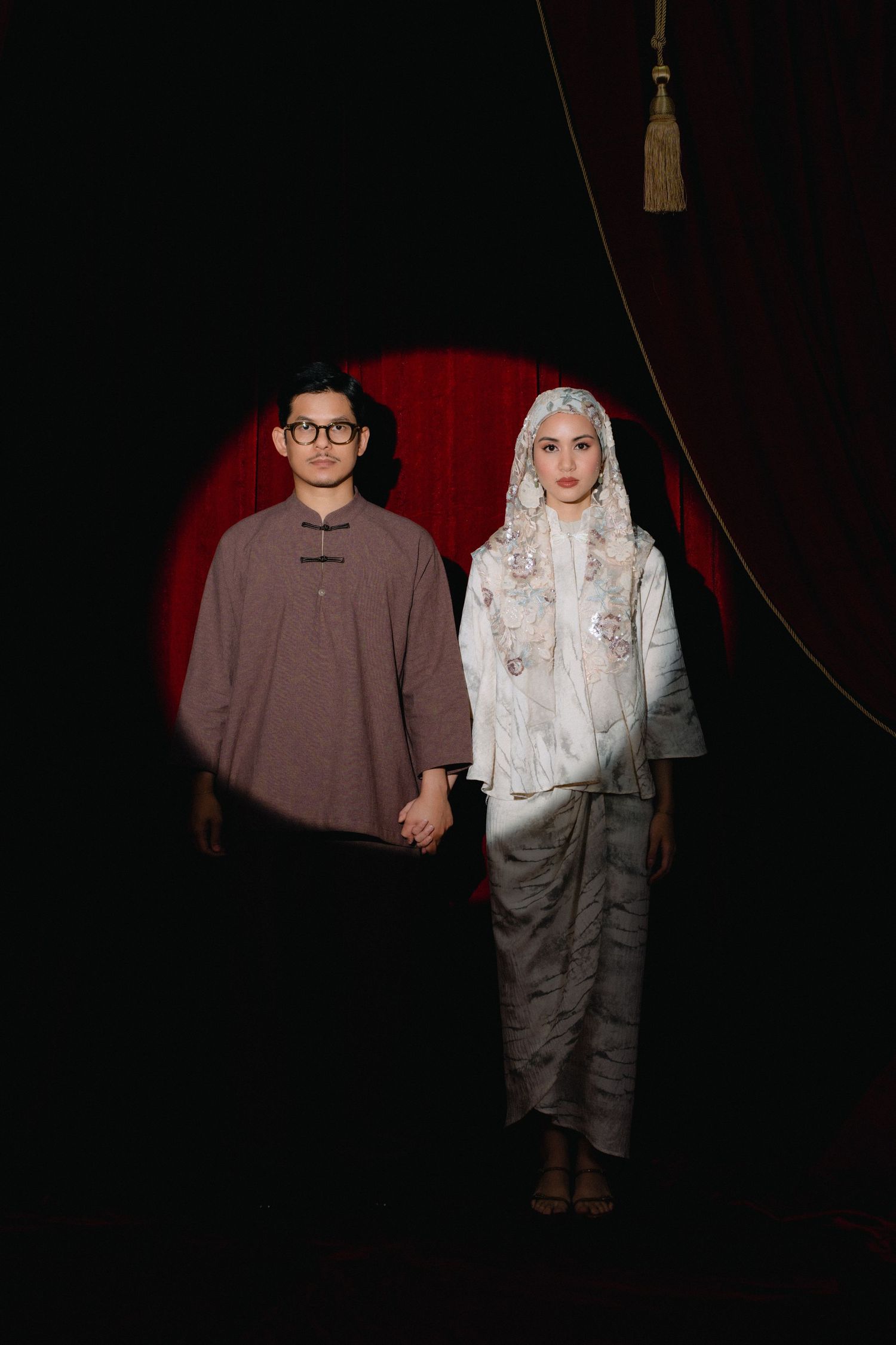 A couple in traditional Muslim attire holds hands against a dramatic dark backdrop with red curtain.