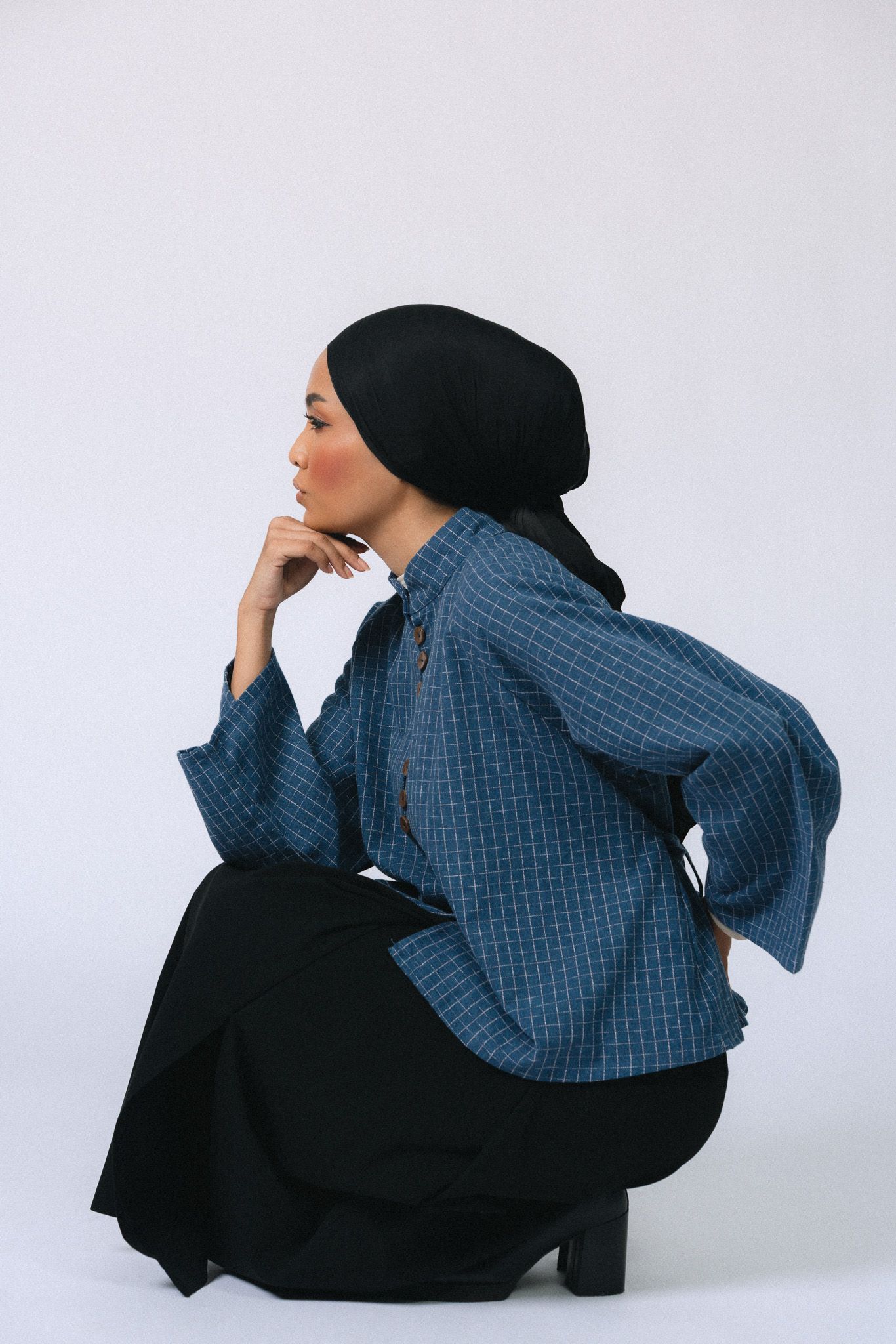 Woman in blue plaid top and black hijab sits thoughtfully, hand on chin, wearing a black skirt on white background.