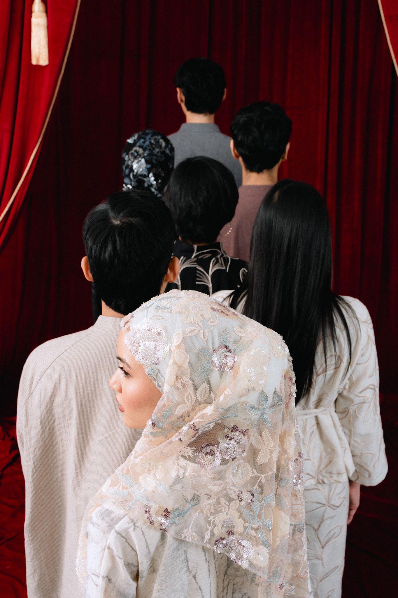 Group of people in traditional attire standing before red curtains, viewed from behind, with one woman in lace hijab.