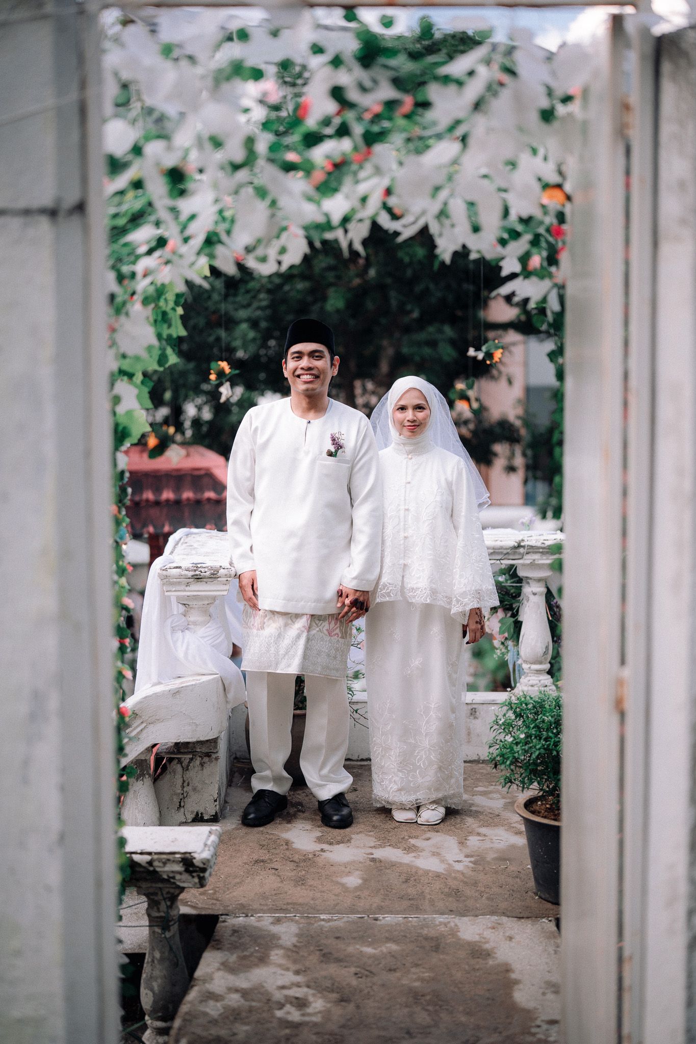 Muslim couple in white traditional wedding attire standing under floral arch outdoors.