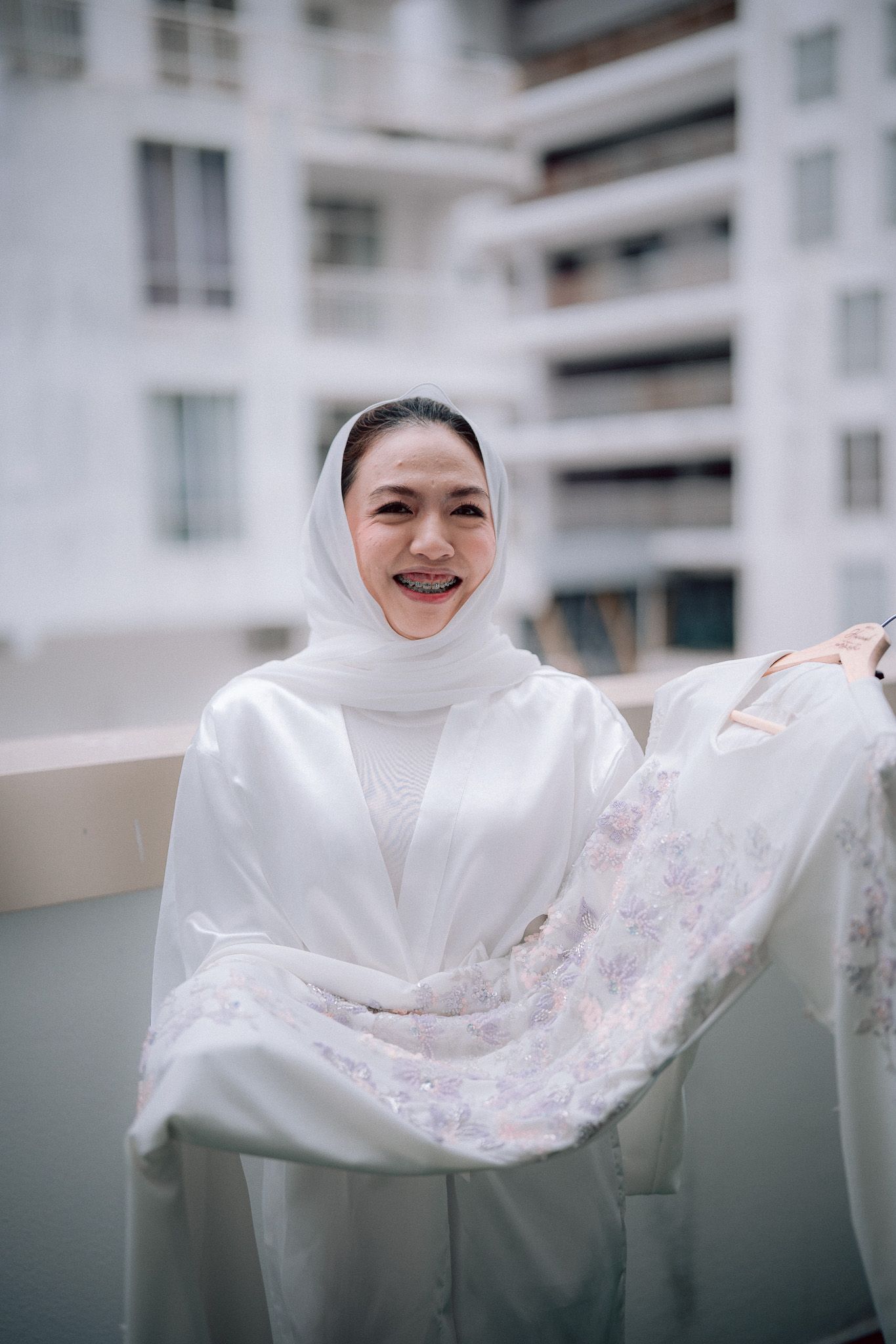 Smiling woman in white hijab and robe holding floral fabric on a balcony with building in background.