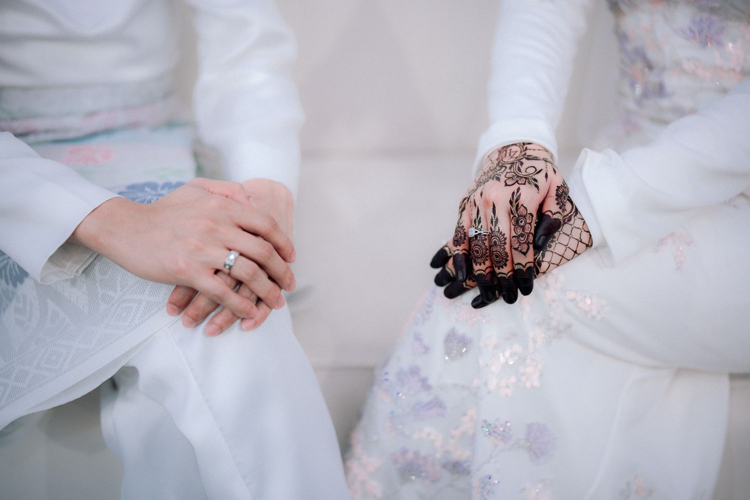 Two women in white attire sit together, one wearing a ring and the other adorned with intricate henna on her hands.