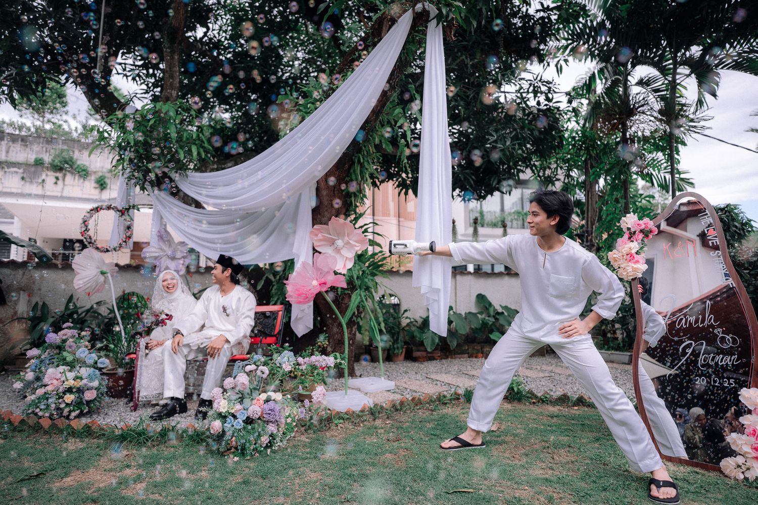 Outdoor garden wedding setup with couple seated among flowers, white fabric draping, and a person posing in white outfit.