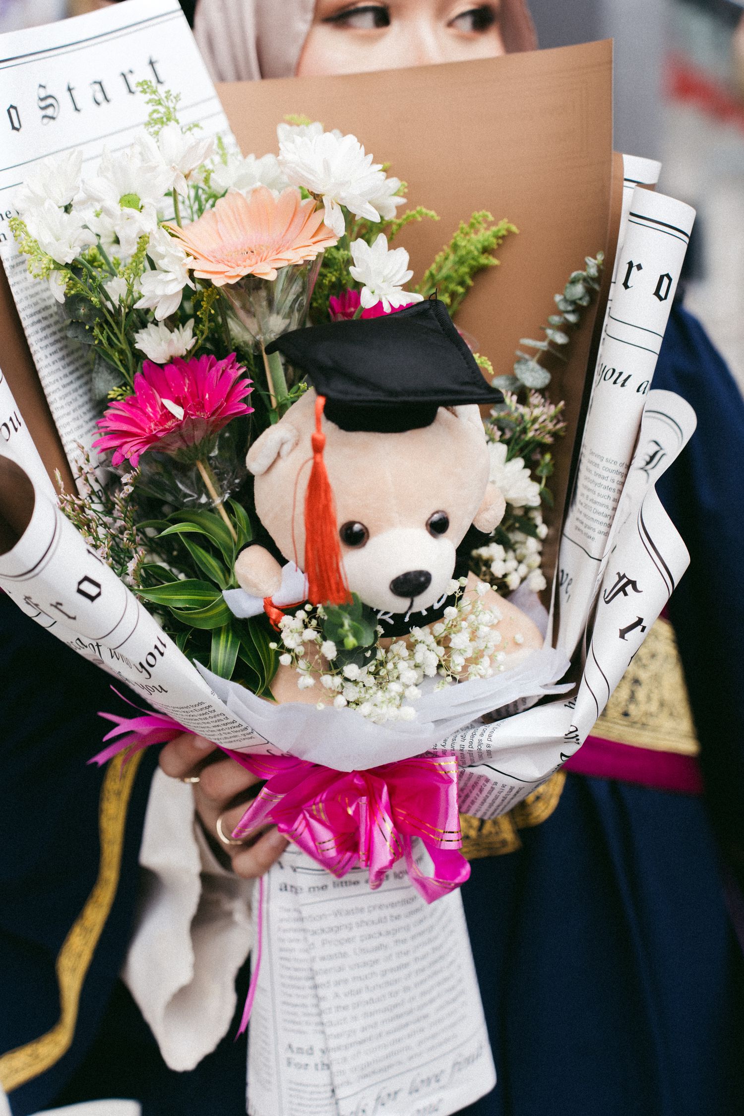 A graduation bouquet featuring white and pink flowers with a plush puppy wearing a graduation cap and academic regalia.