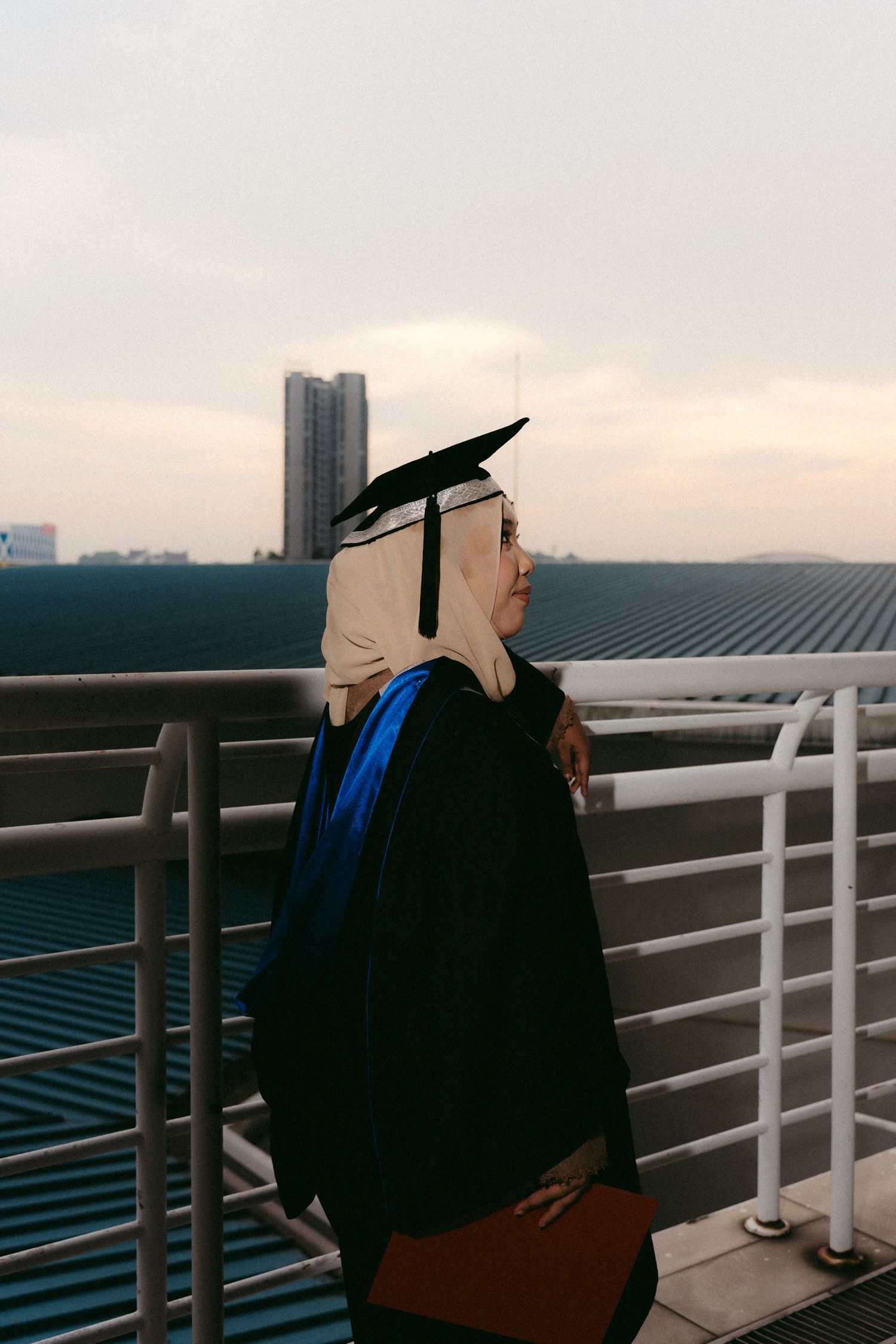 A graduate in black cap and gown with blue stole stands on a rooftop balcony at sunset with city buildings in the background.