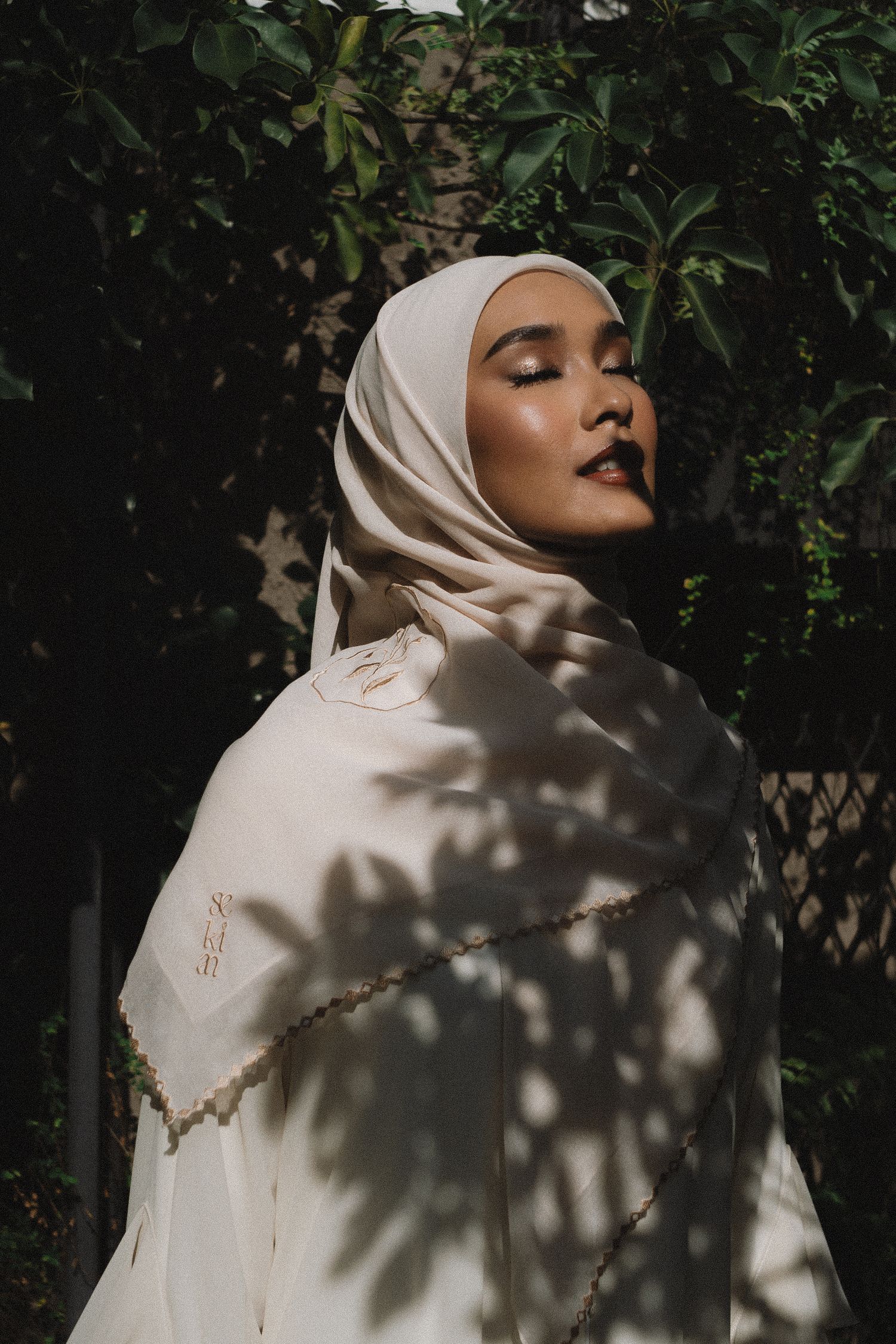 Person in white hijab poses against dark foliage with dramatic shadow patterns.