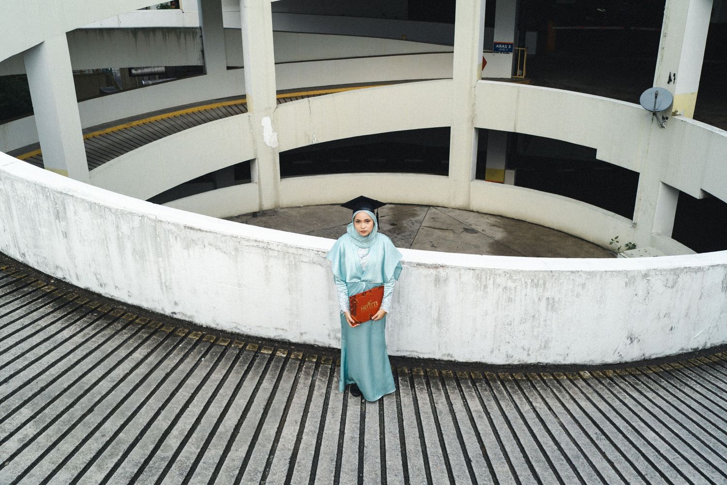 Graduate in light blue outfit stands in curved parking structure with modern architecture.