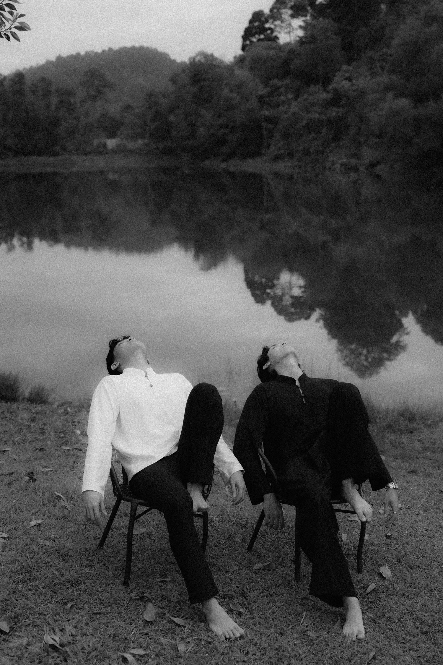 Artistic black and white photo of two people sitting in chairs by lake looking up at sky.