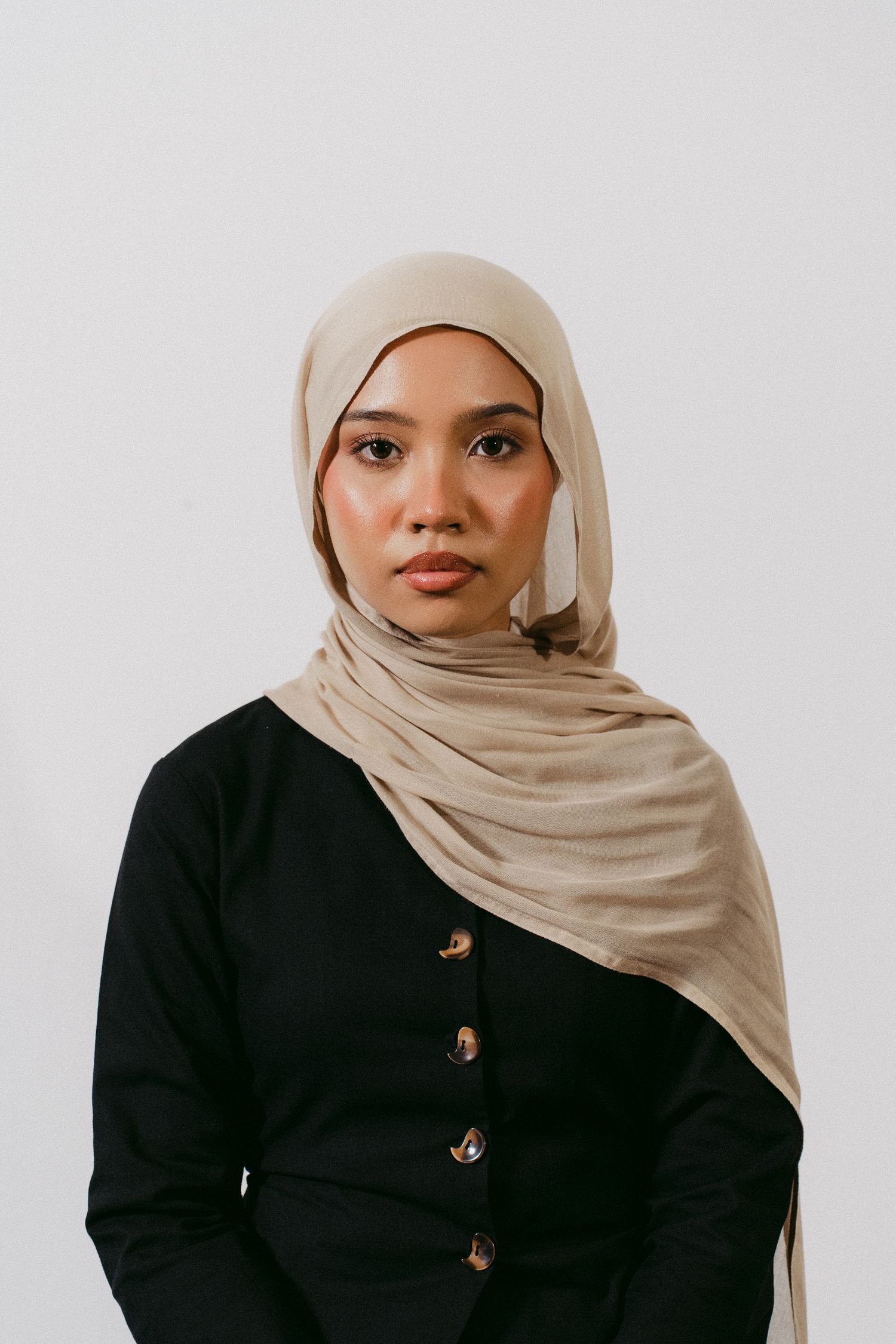 Portrait of woman in beige hijab and black button-up top against white background.