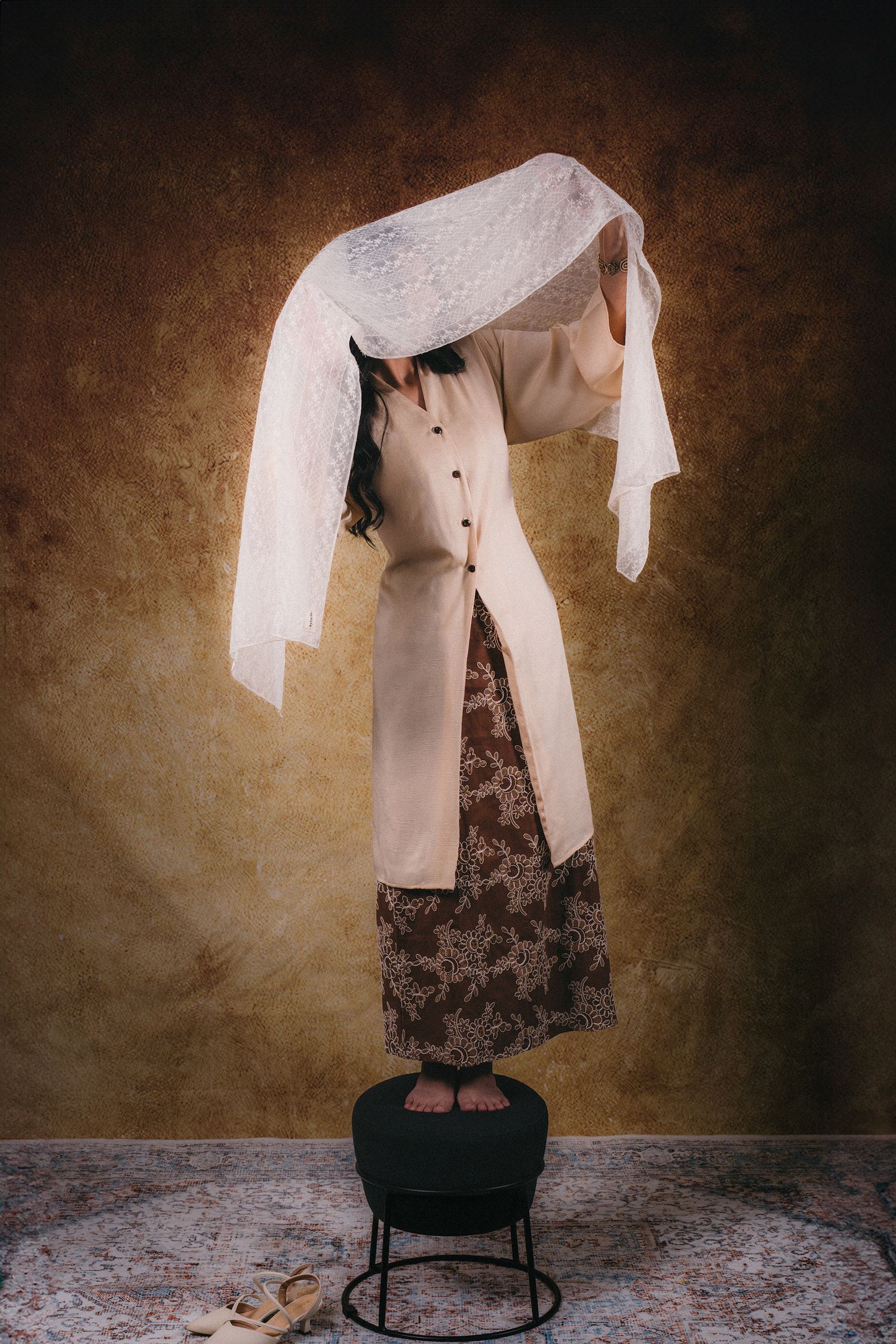 A floating dress display with lace veil suspended above a black stool against a brown textured wall.