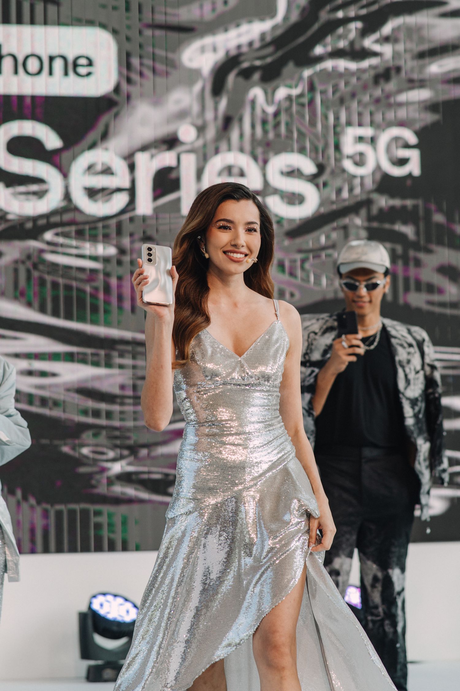 Fashion show features sparkly silver evening gown on runway with tech-themed backdrop.