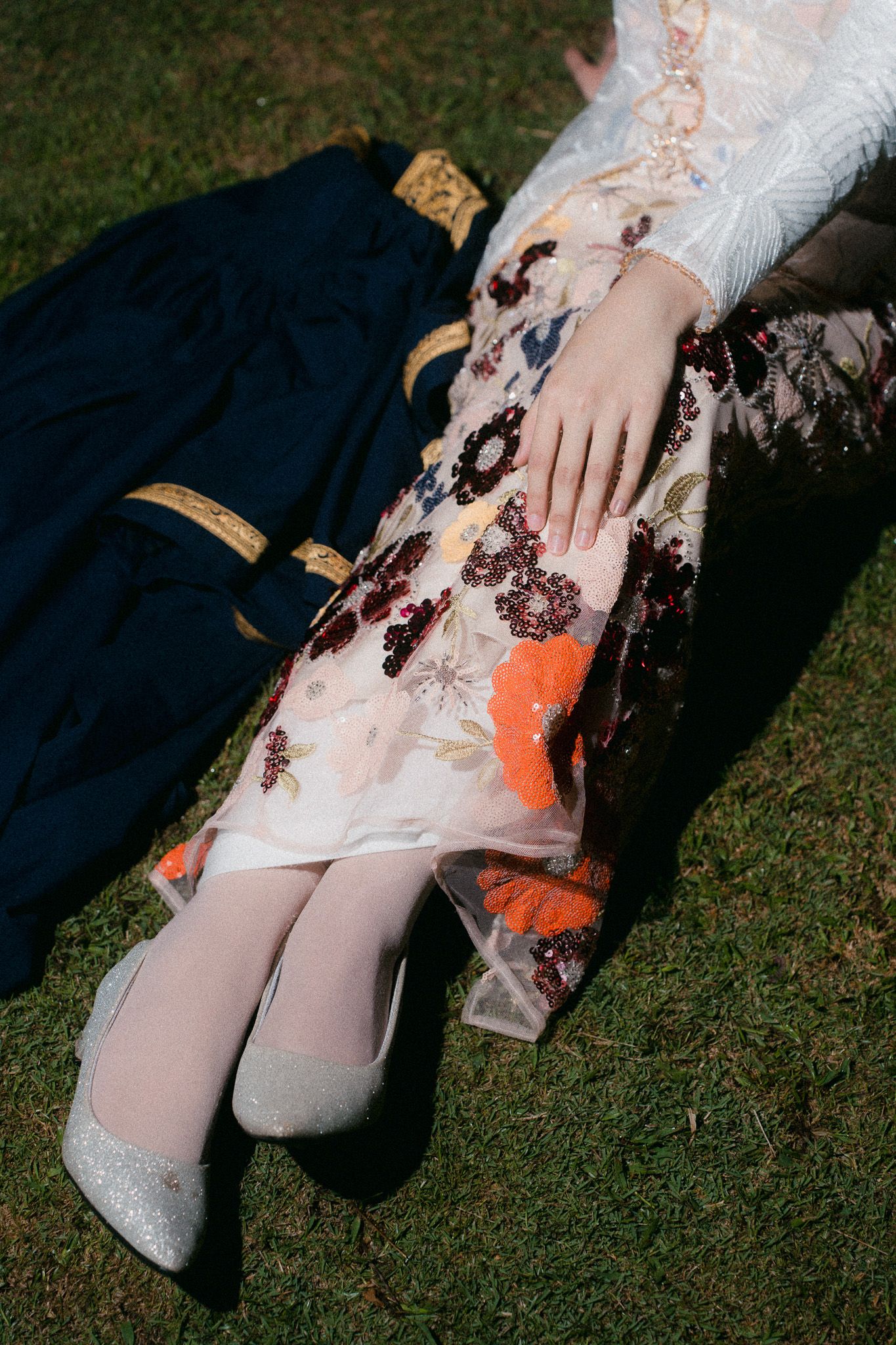 Woman in floral embroidered gown with silver heels sitting on grass beside a dark blue outfit.