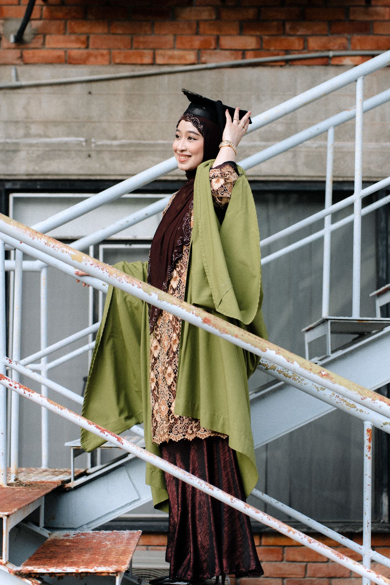 Muslim woman in green abaya and graduation cap smiling on metal stairs outdoors.