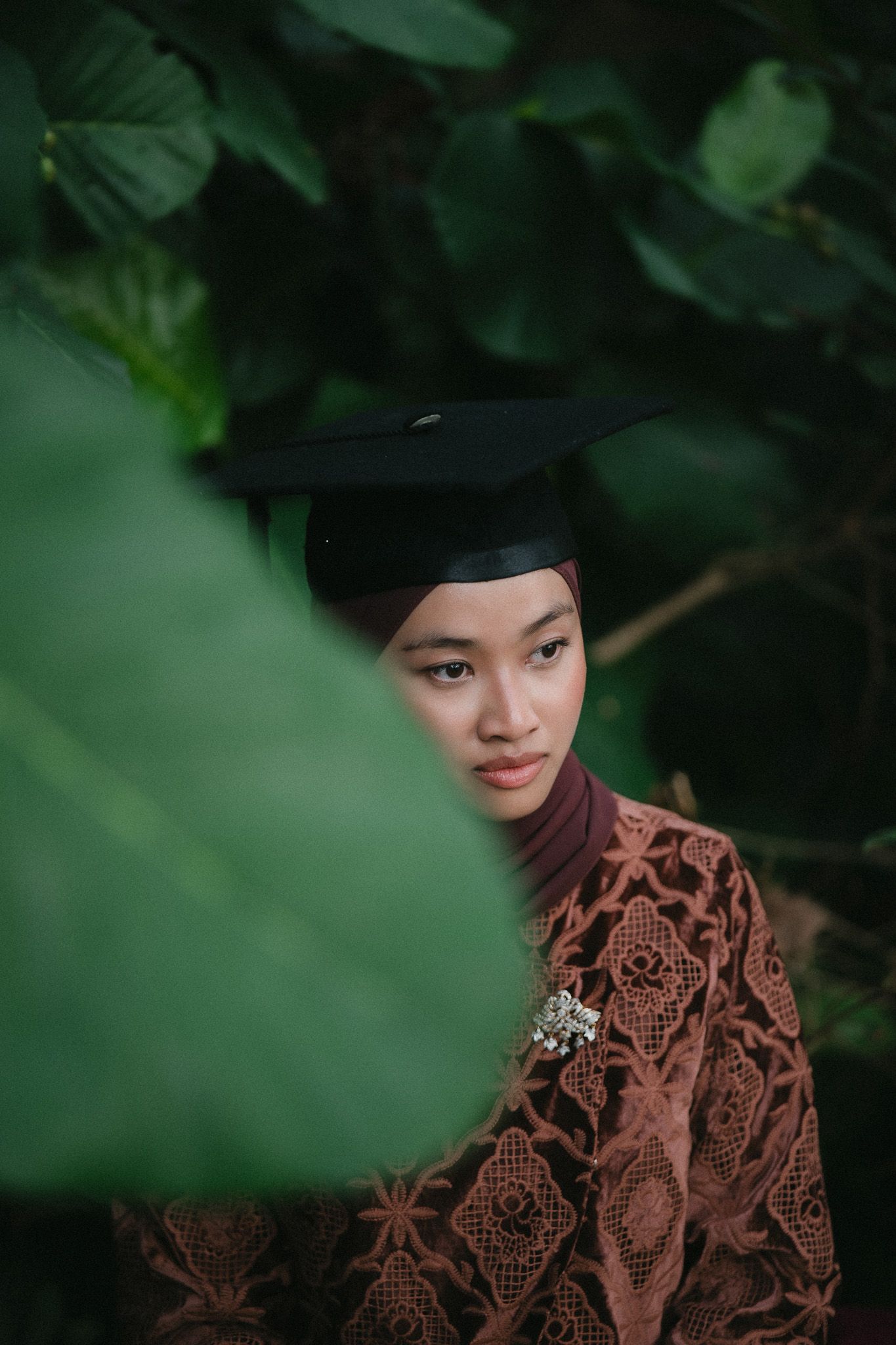 Young woman in hijab and graduation cap poses among lush green tropical leaves wearing brown lace kebaya.