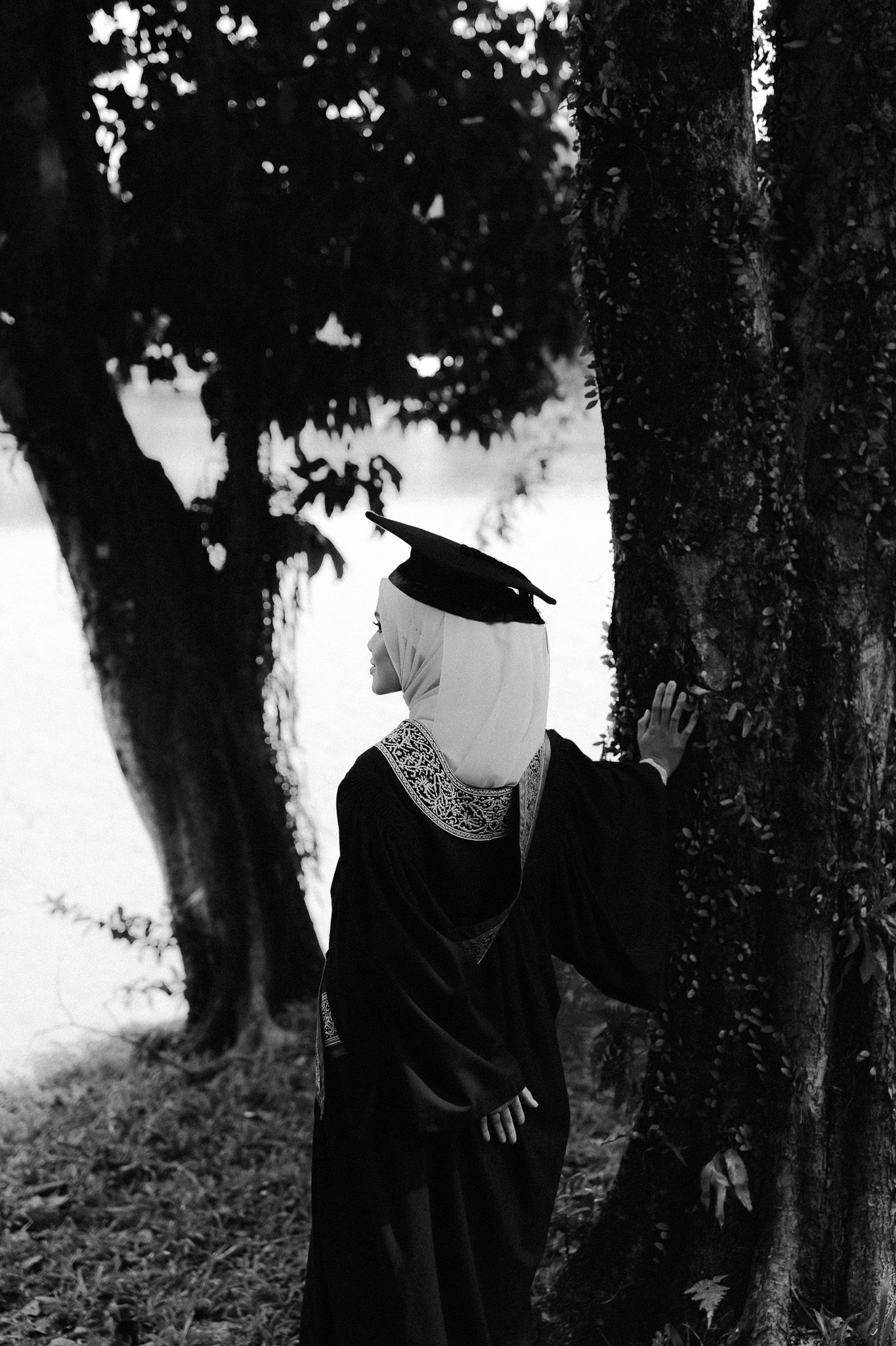 Graduate in black gown and mortarboard cap stands by trees in black and white photo.