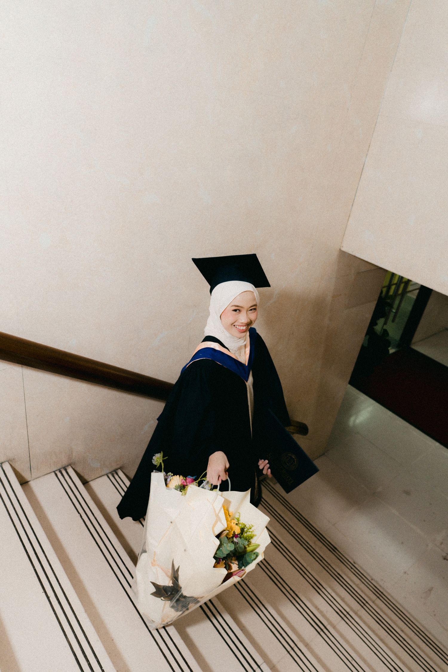 A graduate in cap and gown sits on carpeted stairs holding a bouquet of flowers.