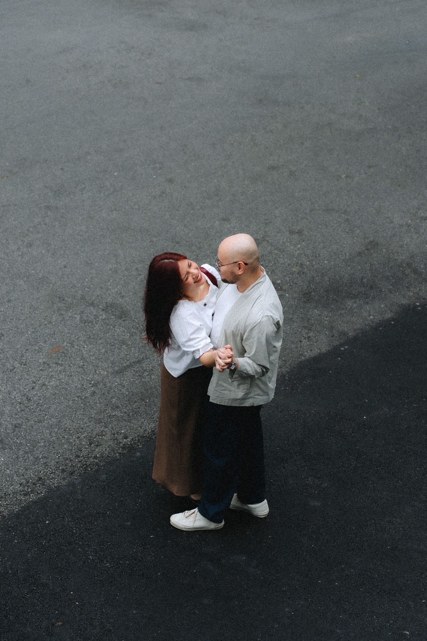 A couple embraces and laughs together while standing on a gray asphalt surface, shot from above.