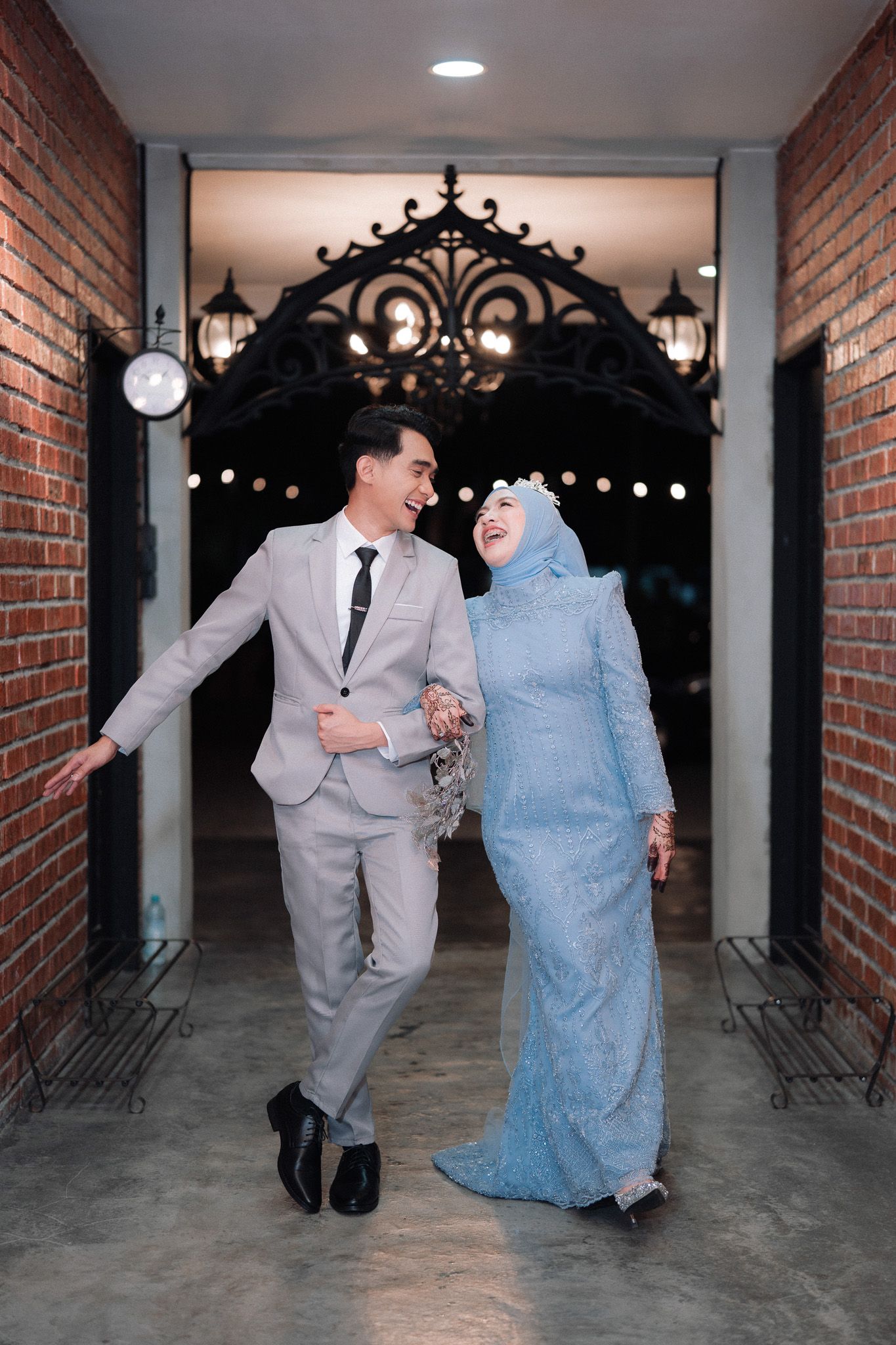 A smiling couple in elegant wedding attire walk together through a decorative archway with brick walls and warm lighting.