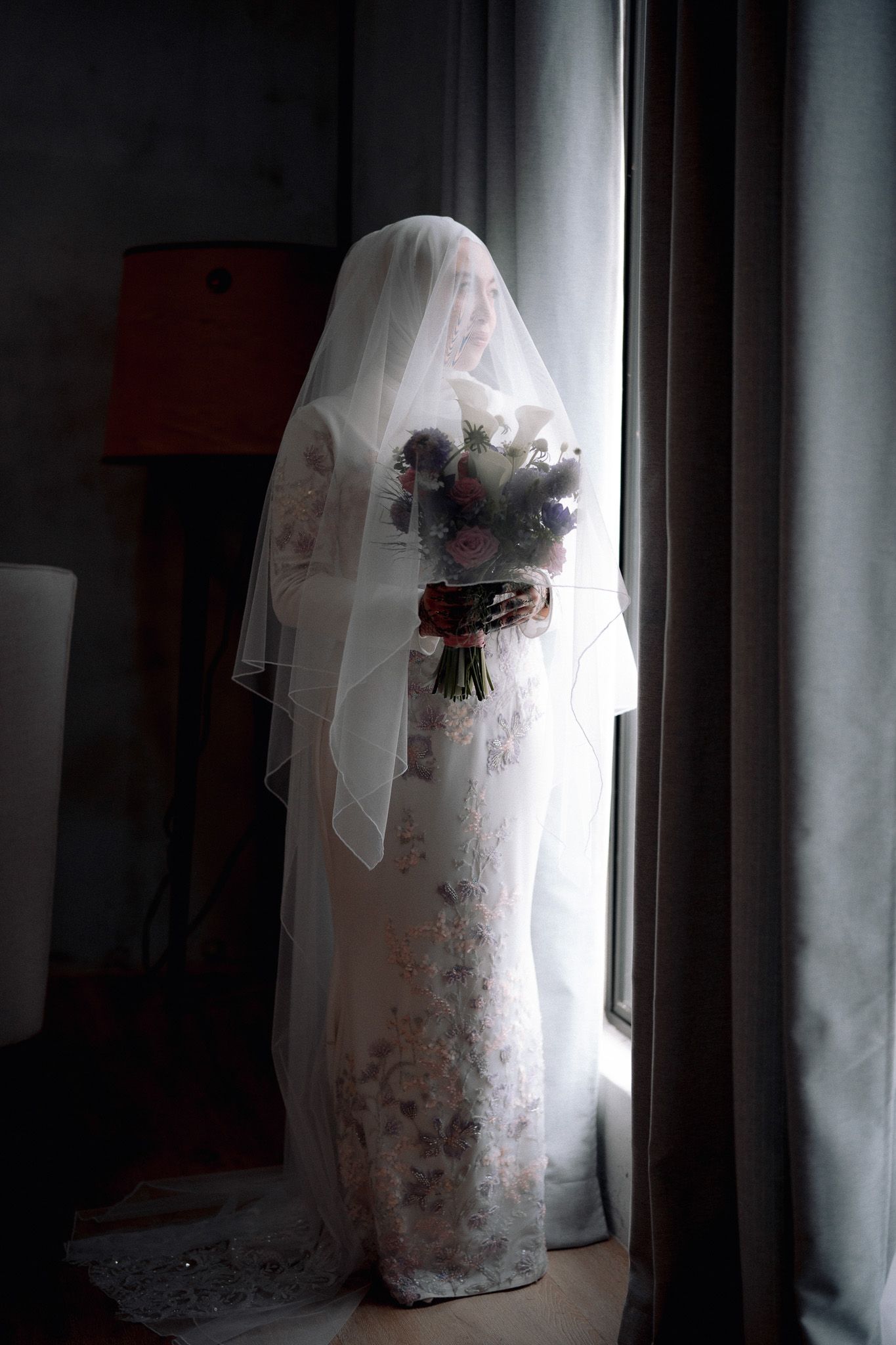 Bride in floral lace gown and white veil holds bouquet of purple and white flowers while standing by a window.
