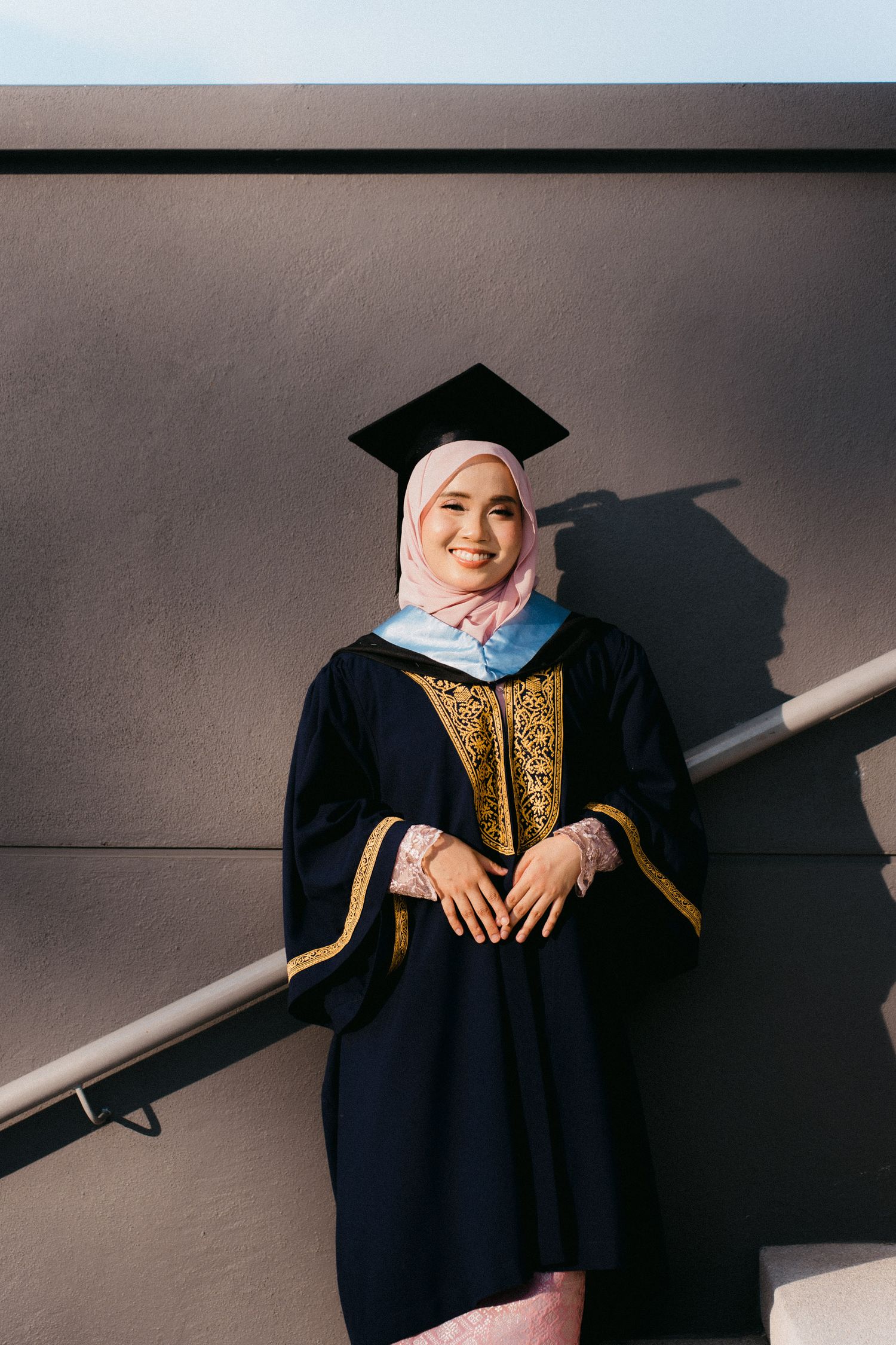 Graduate in traditional black academic gown with gold embellishments poses against grey background in professional photoshoot.