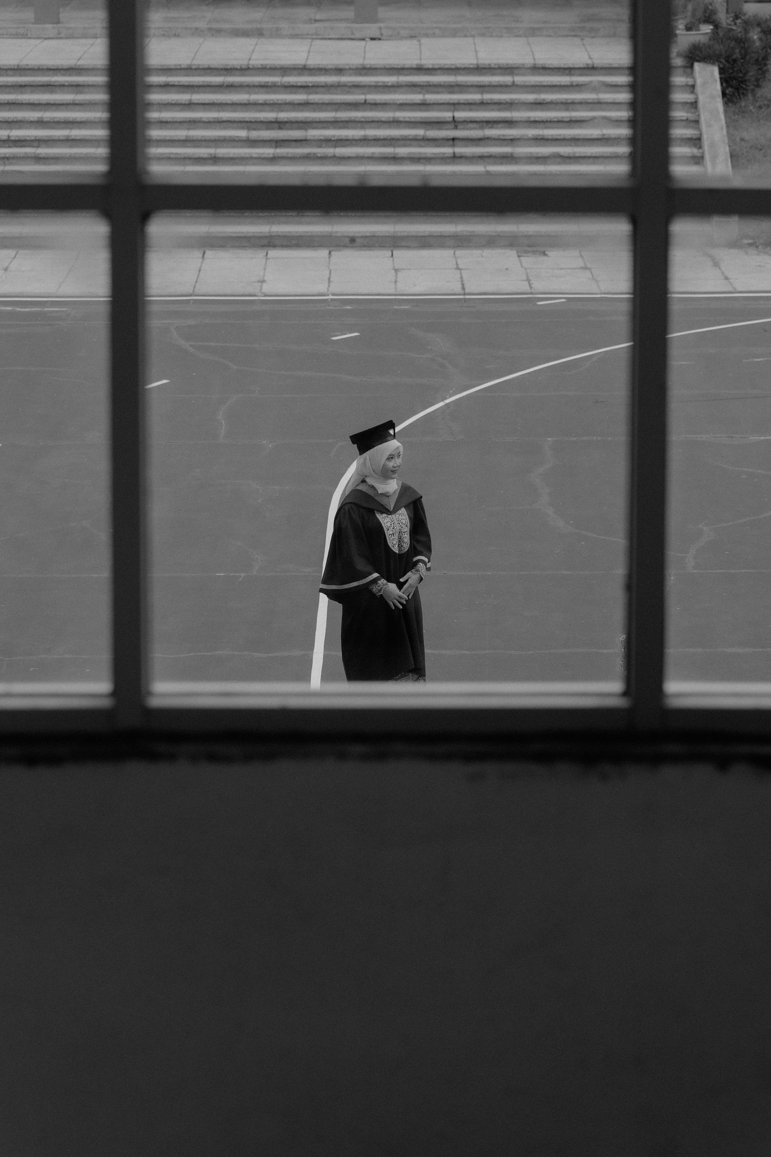 A person in graduation attire stands alone in a gymnasium viewed through window panes creating a grid-like composition.