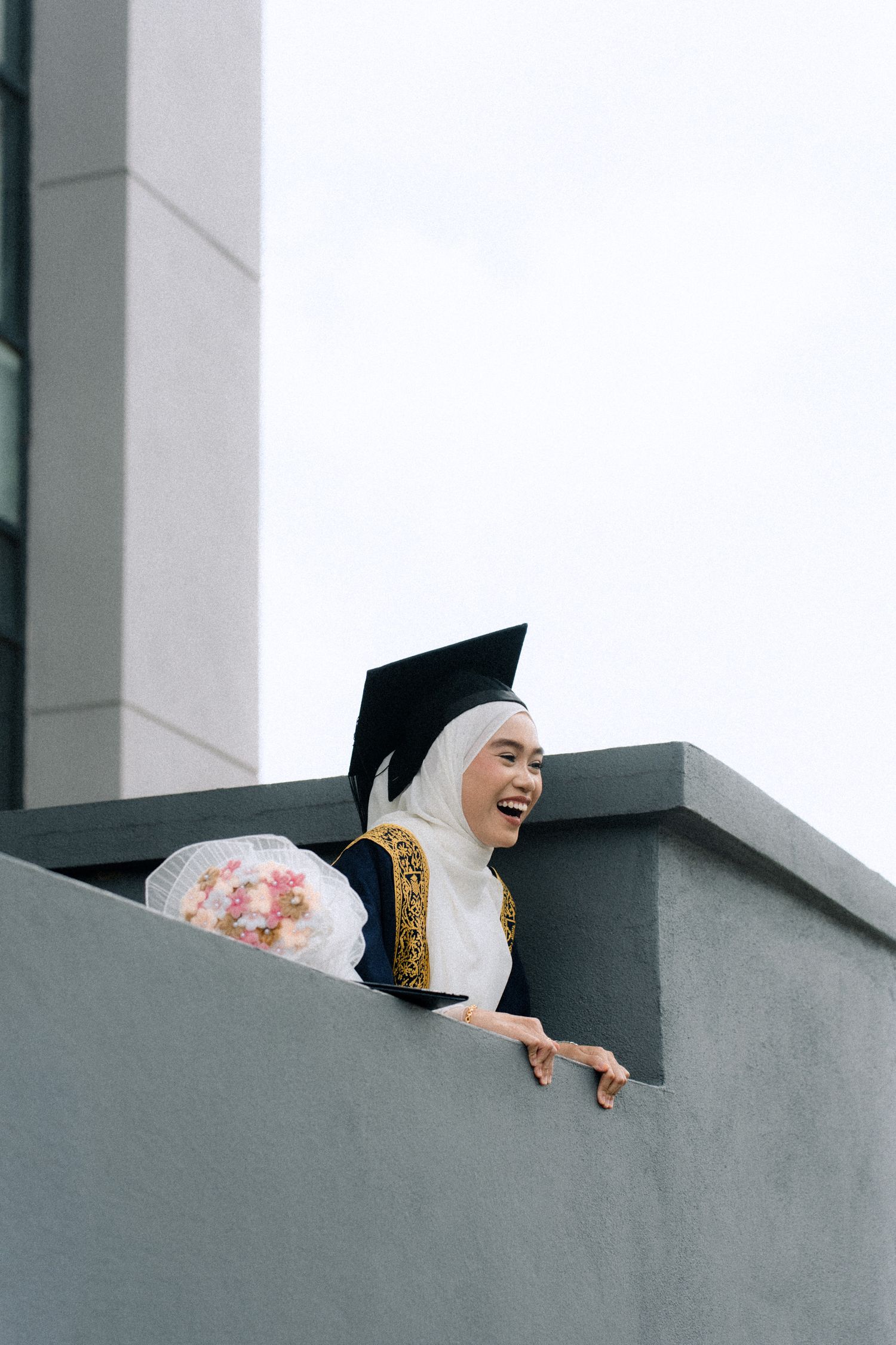 A person in a white hijab and black outfit laughs joyfully while leaning over a gray concrete wall.