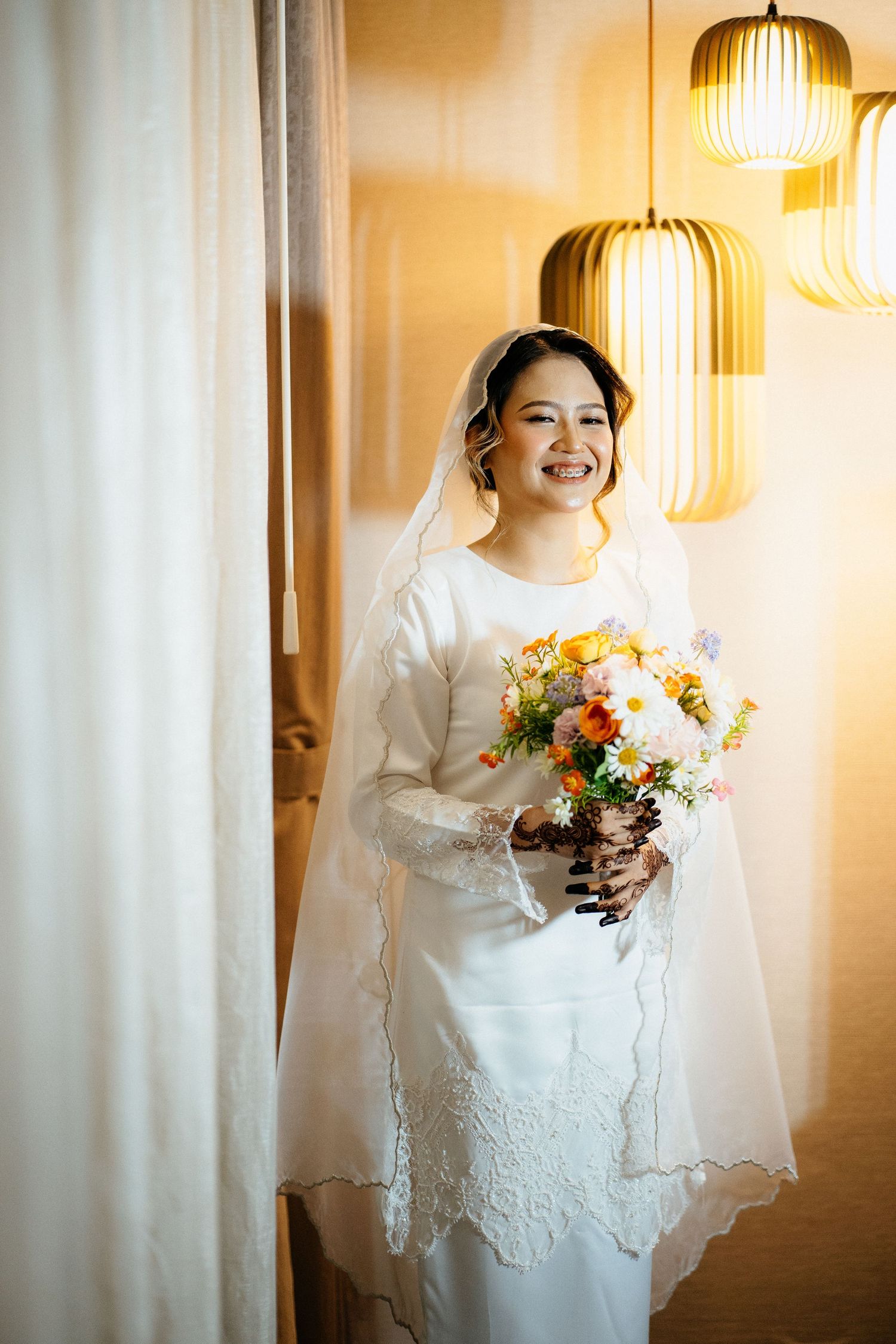 Beautiful bride in white lace dress holds colorful floral bouquet while standing by window under pendant lights.
