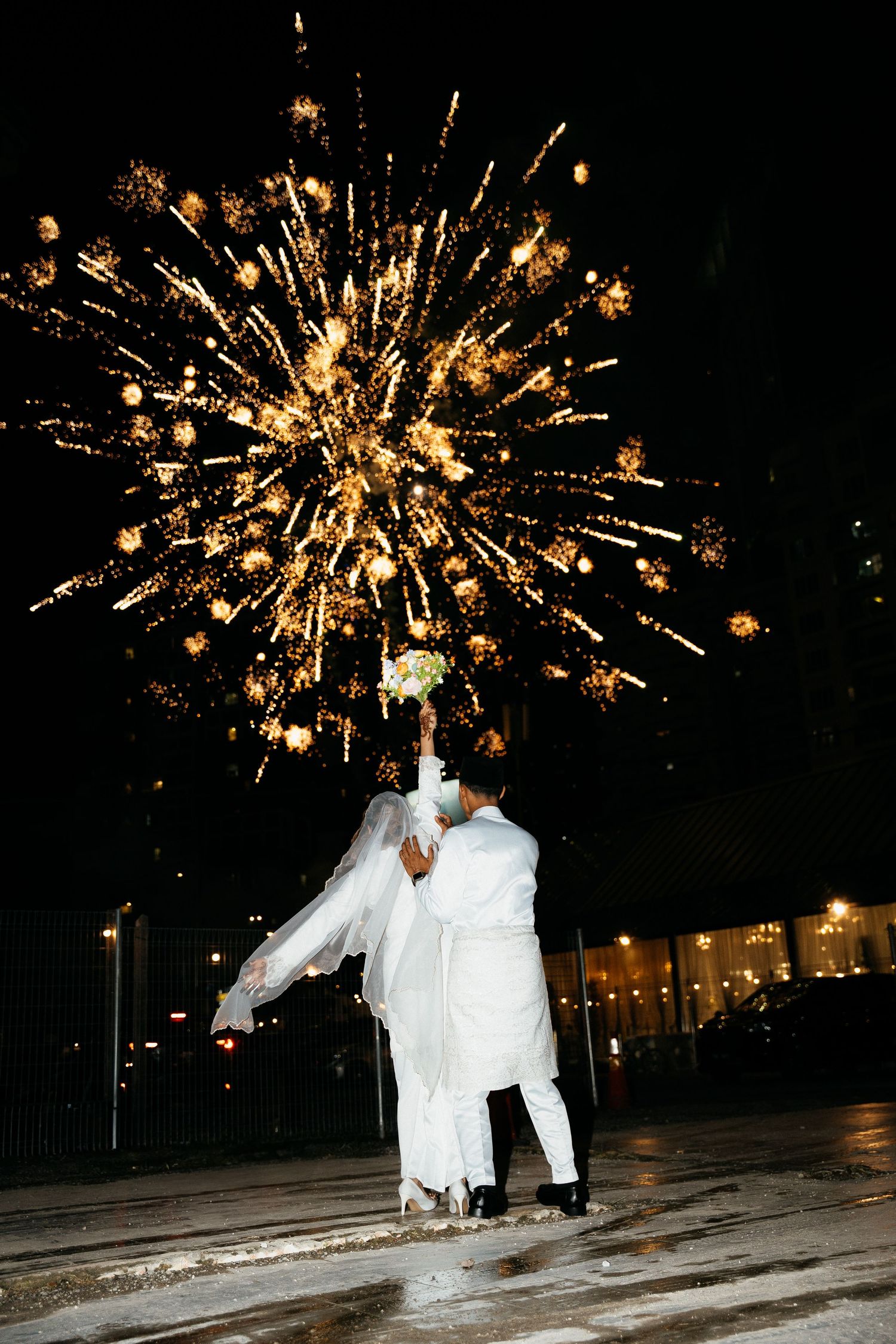 A couple in white embraces under a spectacular golden firework display during a nighttime celebration.
