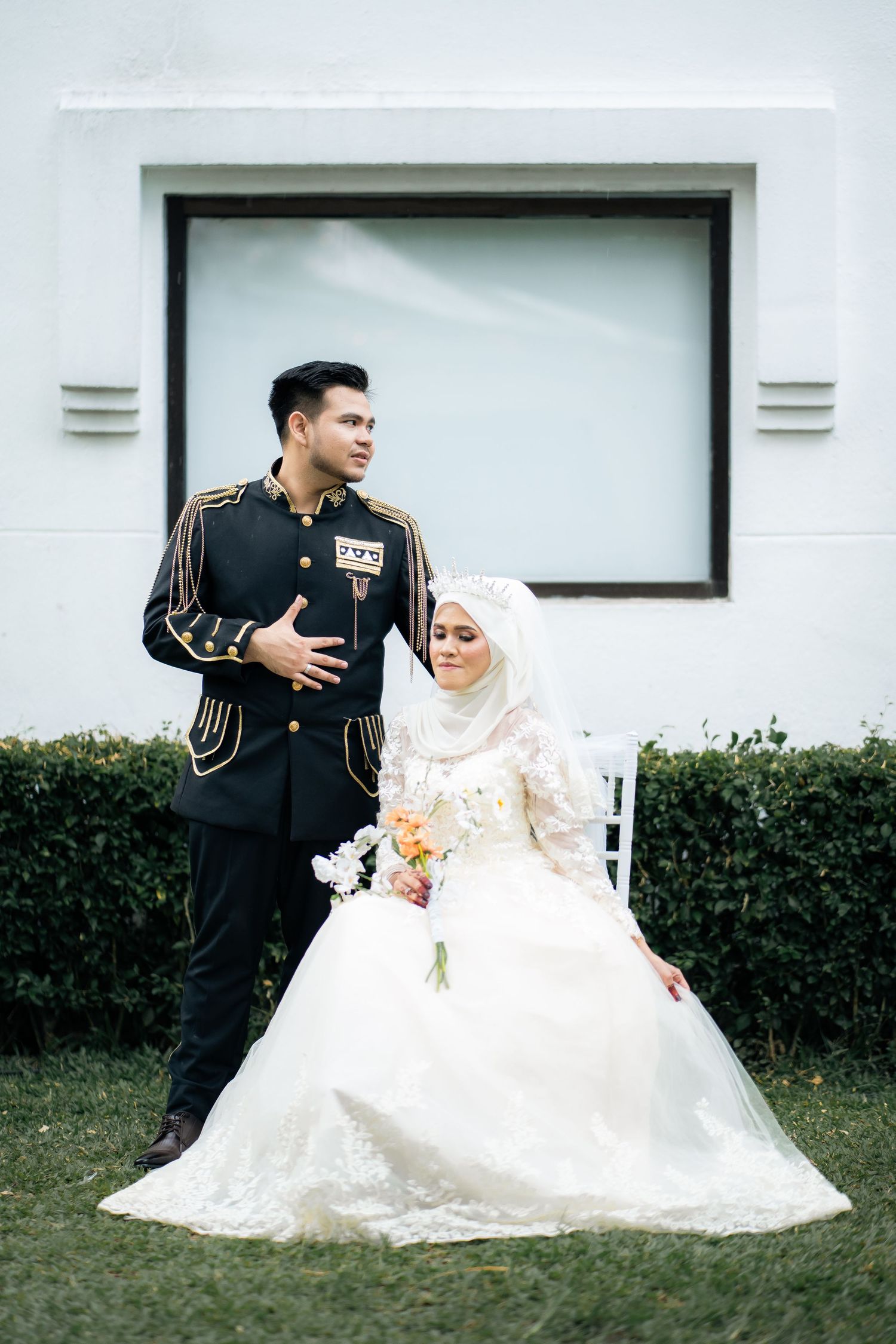 A couple in wedding attire sits together on grass beside white building with hedges.