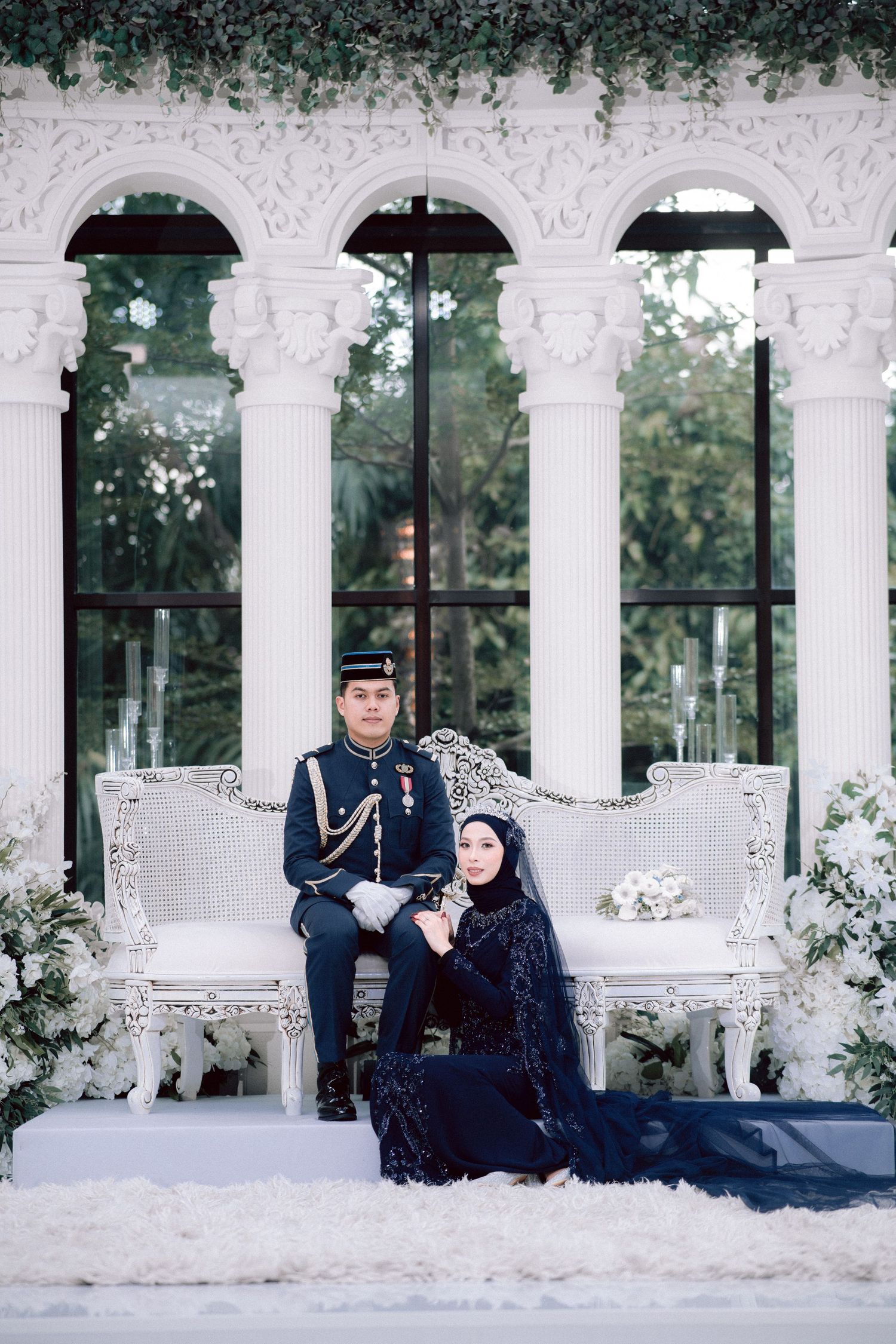 A couple in formal winter attire pose on an ornate white bench surrounded by elegant arches and greenery.