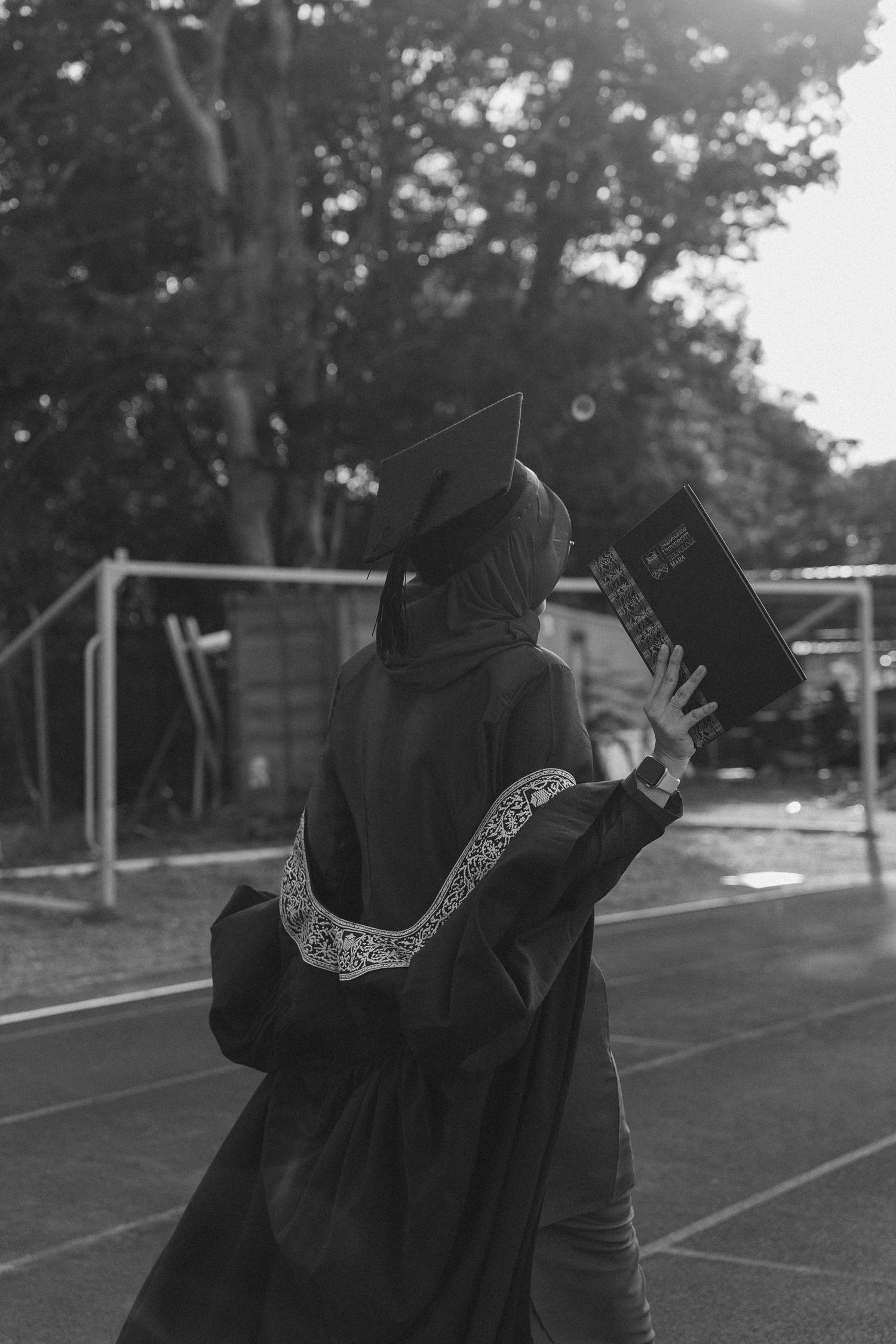 Black and white photo of graduate in cap and gown holding diploma on outdoor track.