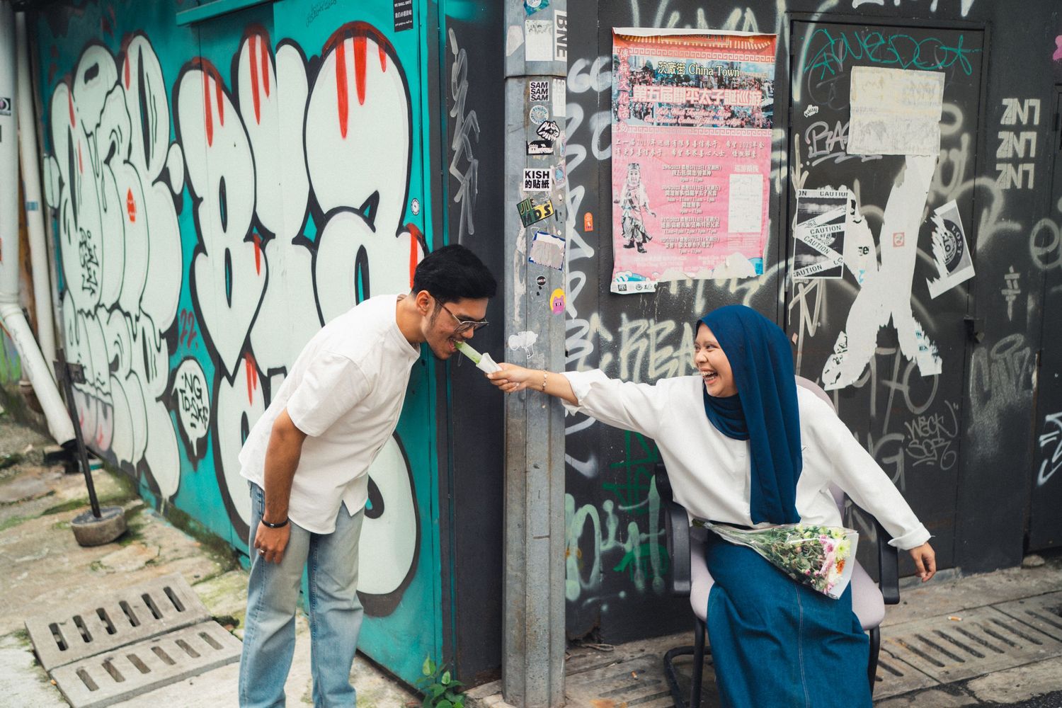 Two people playfully pose in front of a graffiti-covered wall in an urban setting.