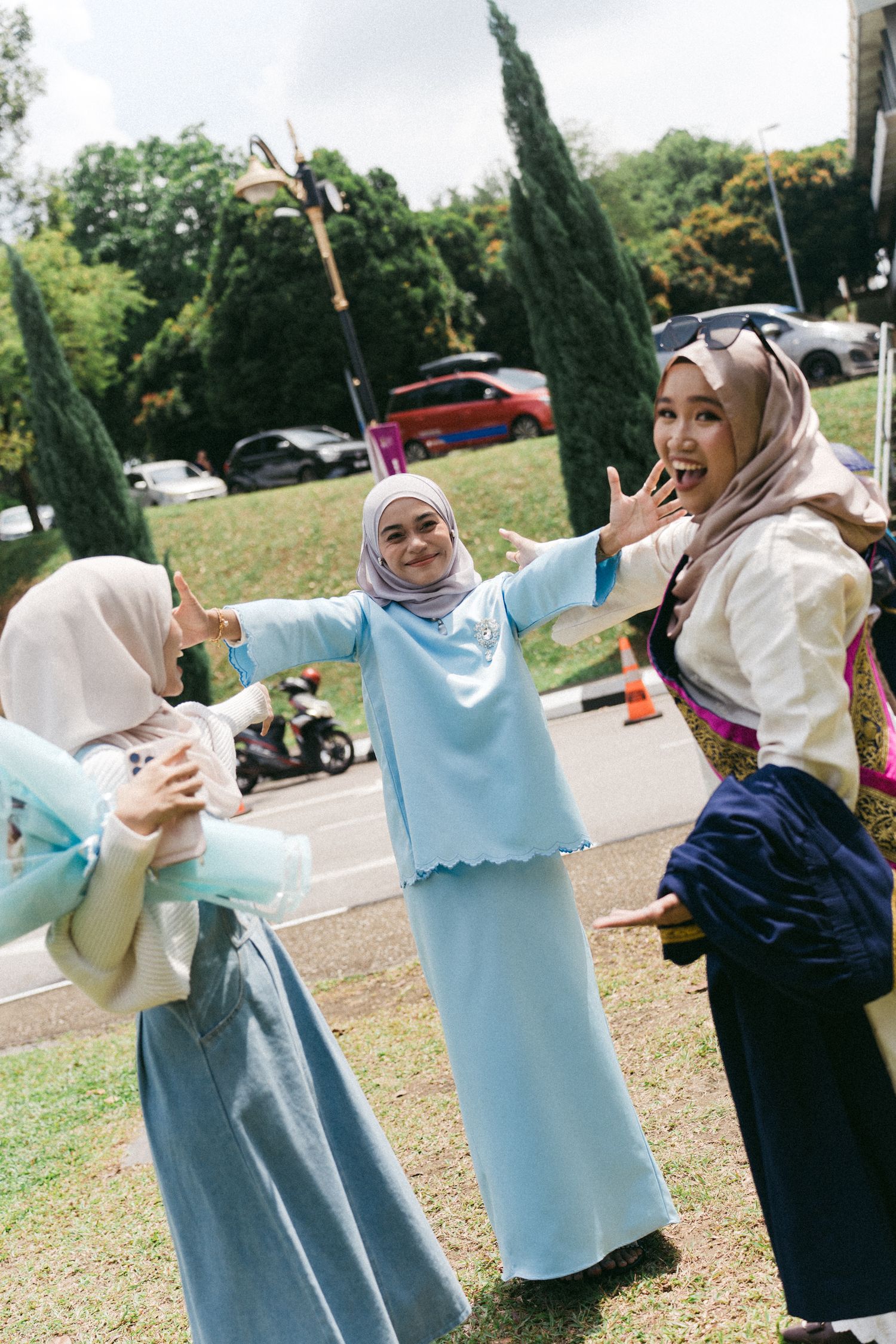 Three friends wearing hijabs share a joyful moment together outdoors near cypress trees and parked cars.