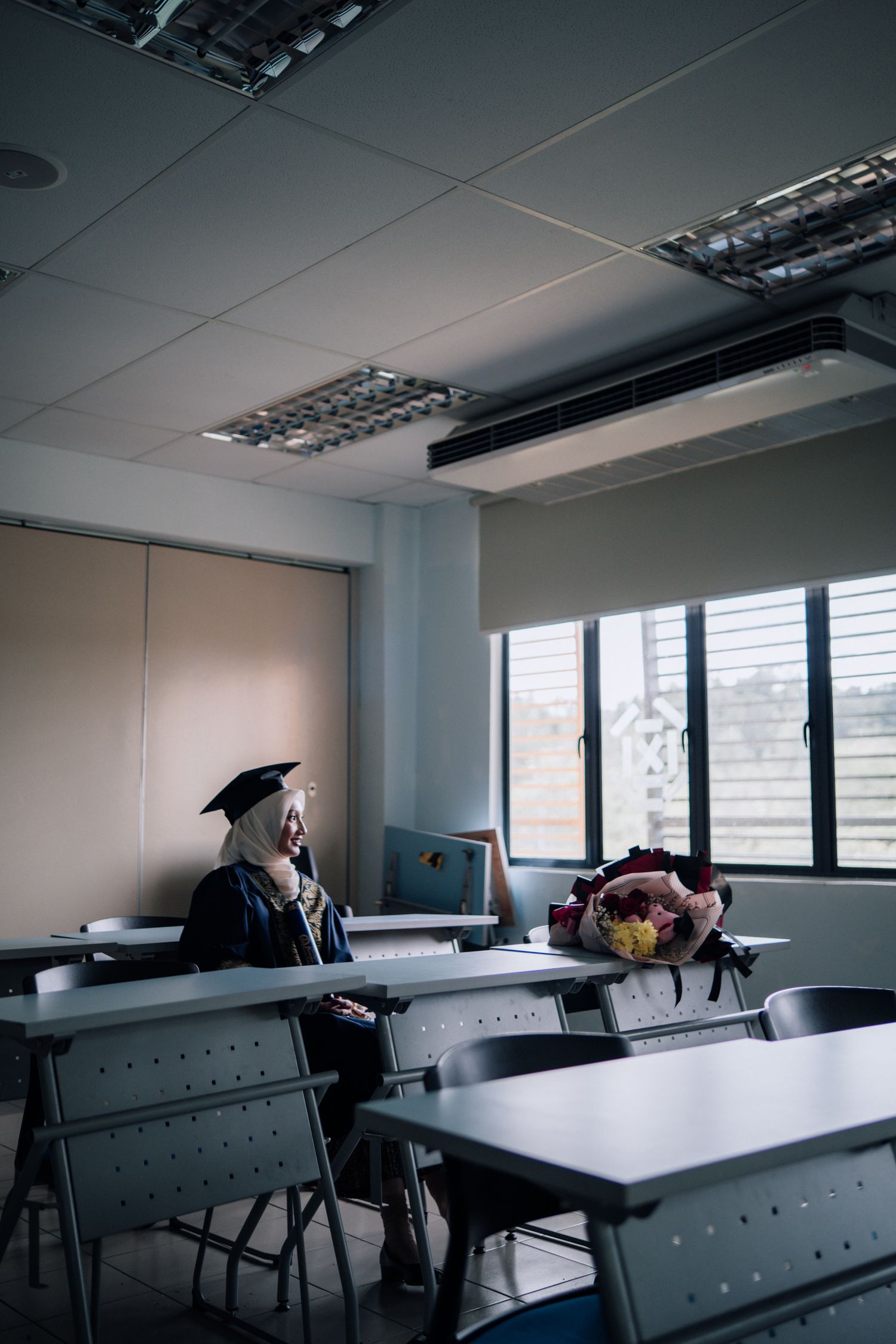 Graduate in cap and gown sits alone in empty classroom with flower bouquet on desk.